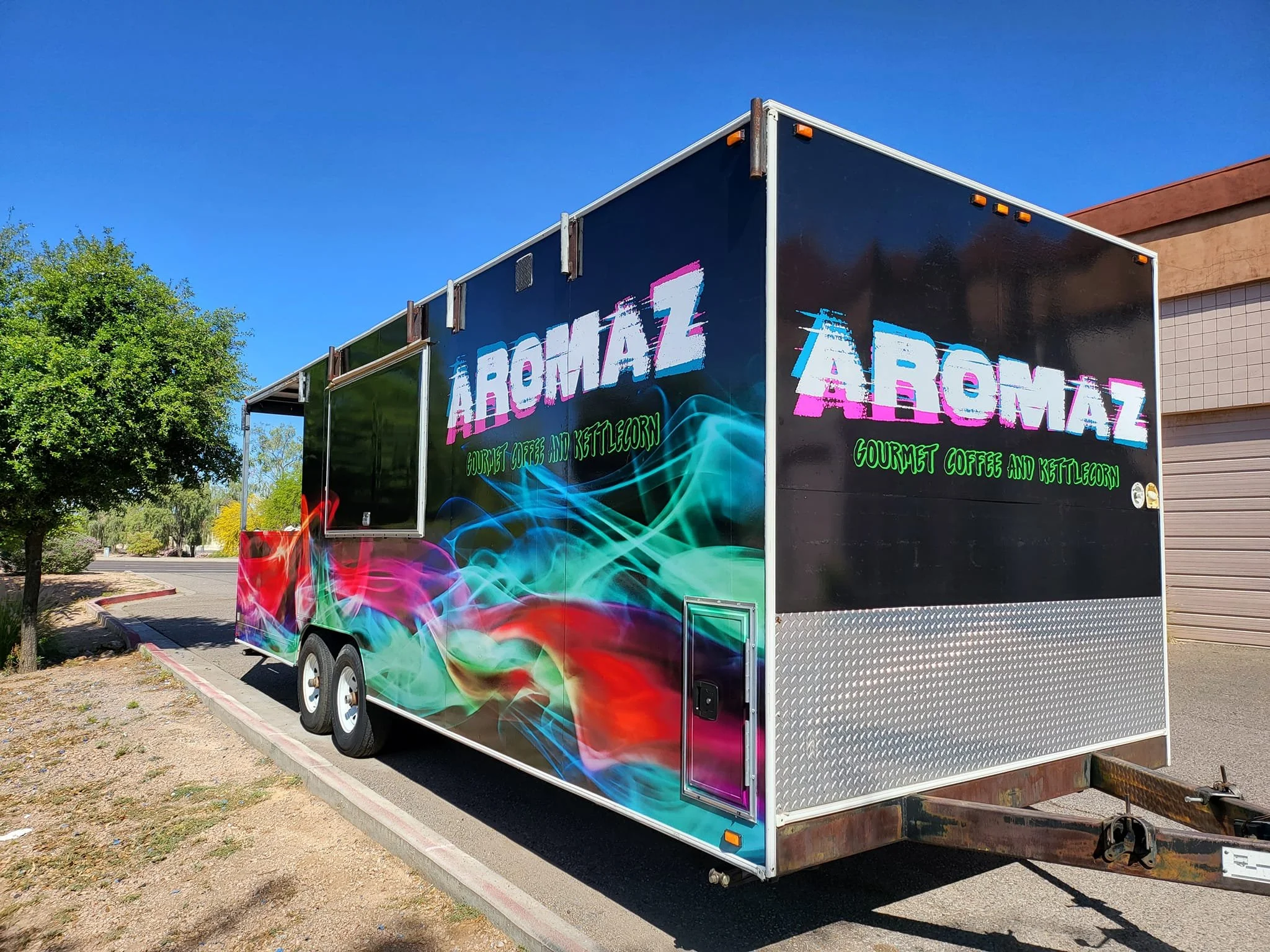 A food truck named 'AROMAZ' offering gourmet coffee and kettlecorn, with a colorful smoke design on the side, parked in a parking lot under a clear blue sky.