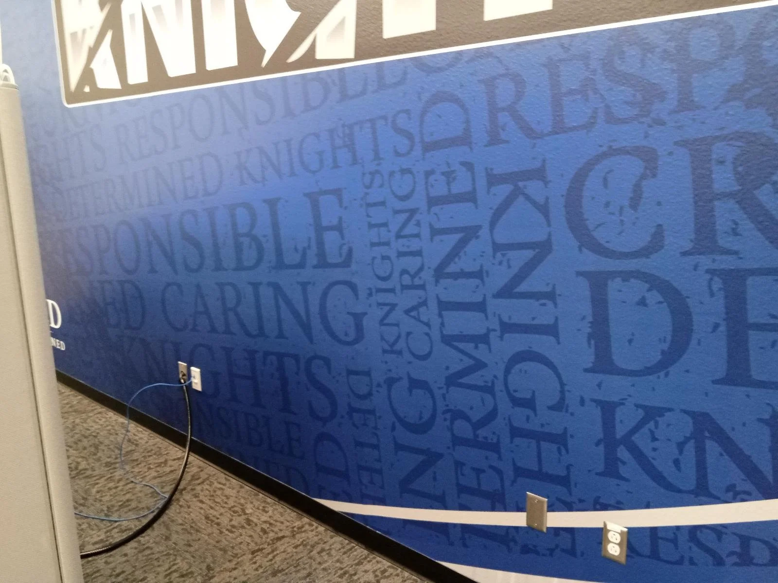 A blue wall with text in big letters and words like 'Responsibility,' 'Caring,' 'Vision,' and 'Trust.' Part of a white framed sign or poster is visible at the top. An electrical outlet and a black cable are on the bottom left.