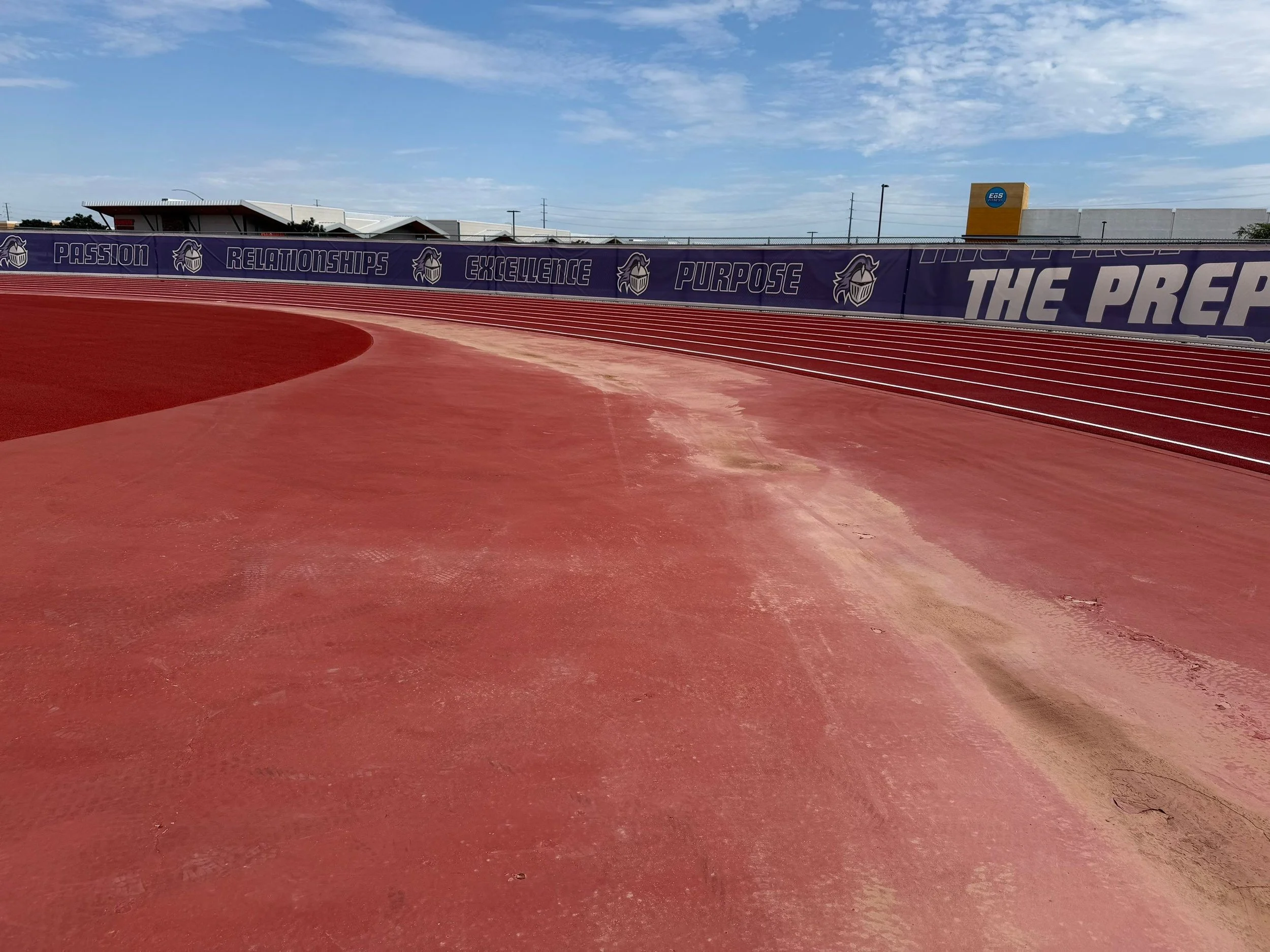 An outdoor athletic track with red surface and white lane markings, surrounded by a purple fence displaying motivational words and a logo of a lion wearing a mask. The sky is partly cloudy.