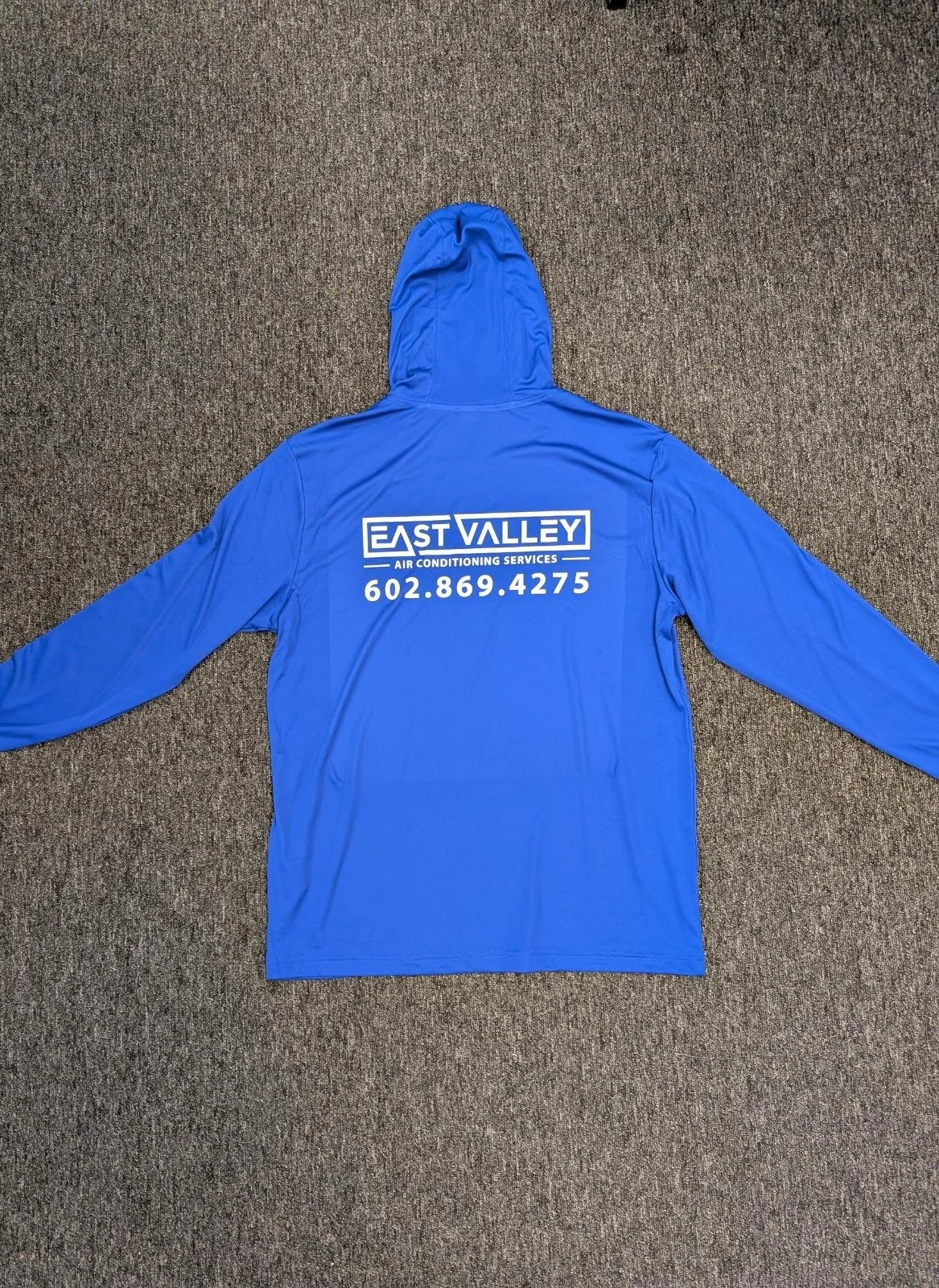 Blue hooded jacket with logo and contact information for East Valley Air Conditioning Services on the back, lying flat on a brown carpet.