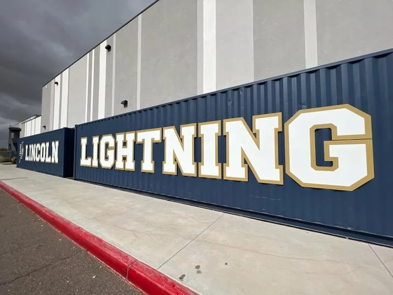 A large blue shipping container with the words 'LINCOLN LIGHTNING' in big white and gold letterings, placed outside a building with a concrete sidewalk and curb.