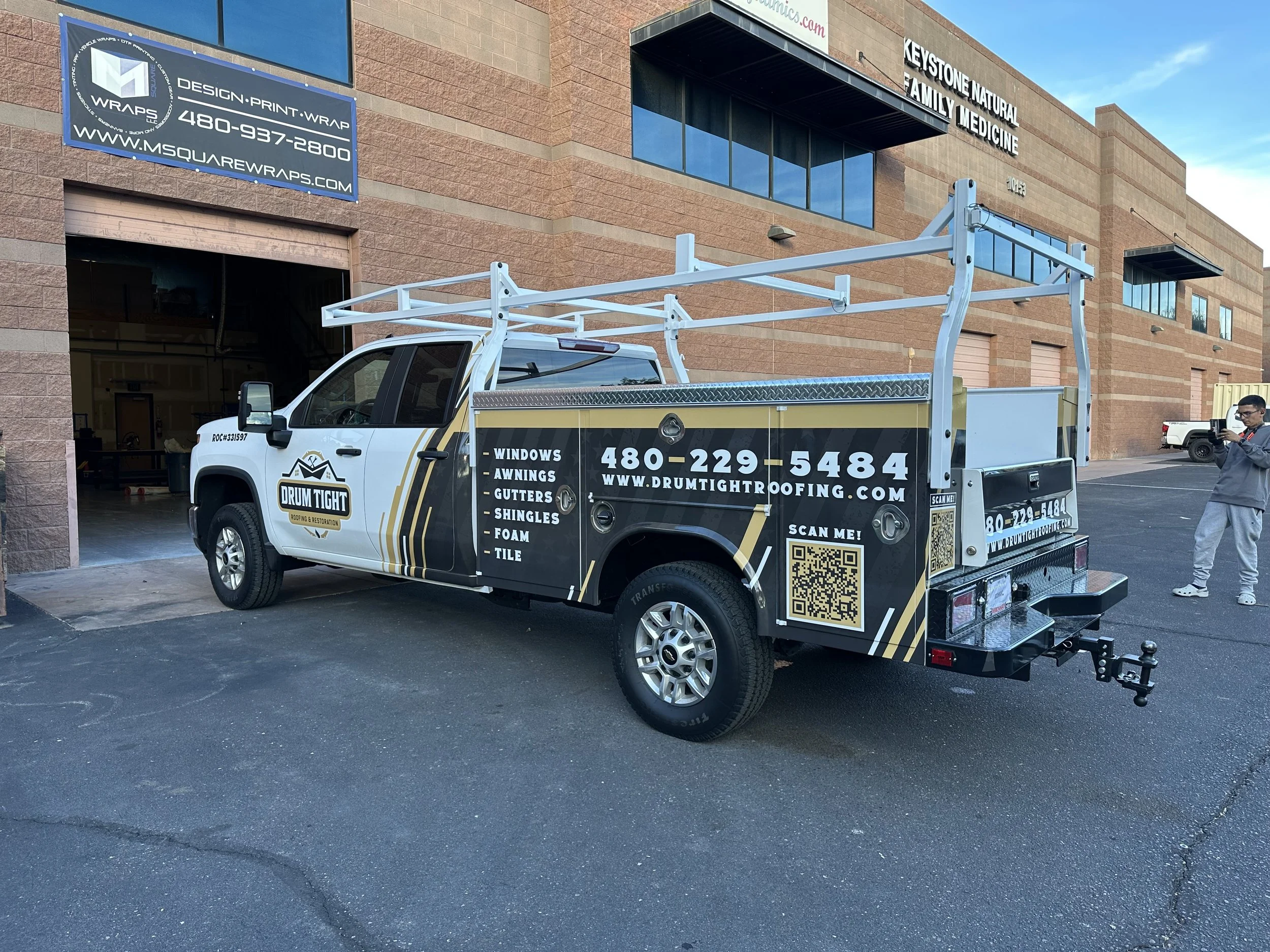 A white work truck with branding for Drum Tight Roofing & Restoration, parked in front of a commercial building. The truck has a ladder rack and signage listing services such as windows, awnings, gutters, shingles, foam, and tile, along with contact 