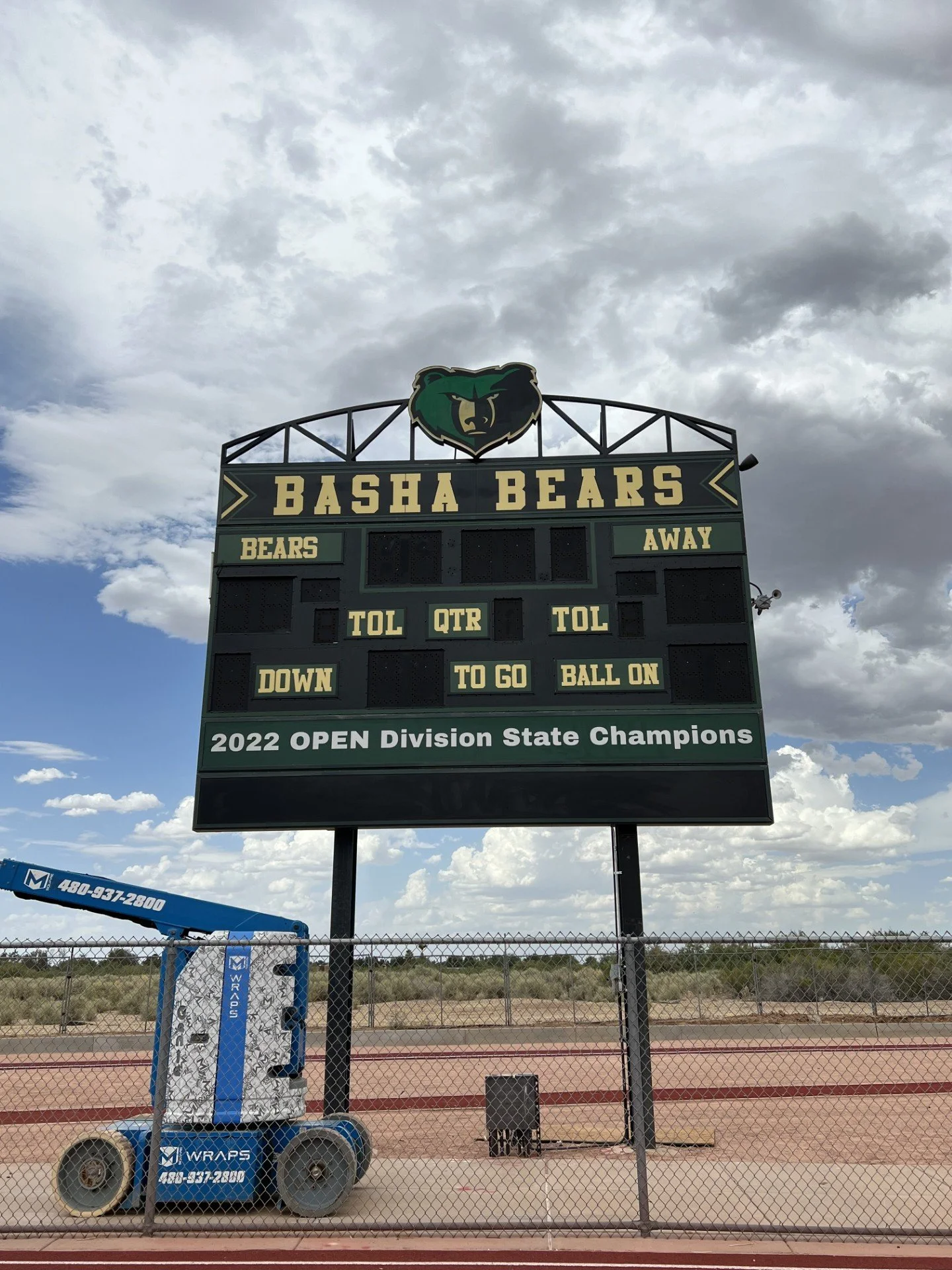 Football scoreboard at Basha Bears stadium showing team info and championship title with cloudy sky in background.
