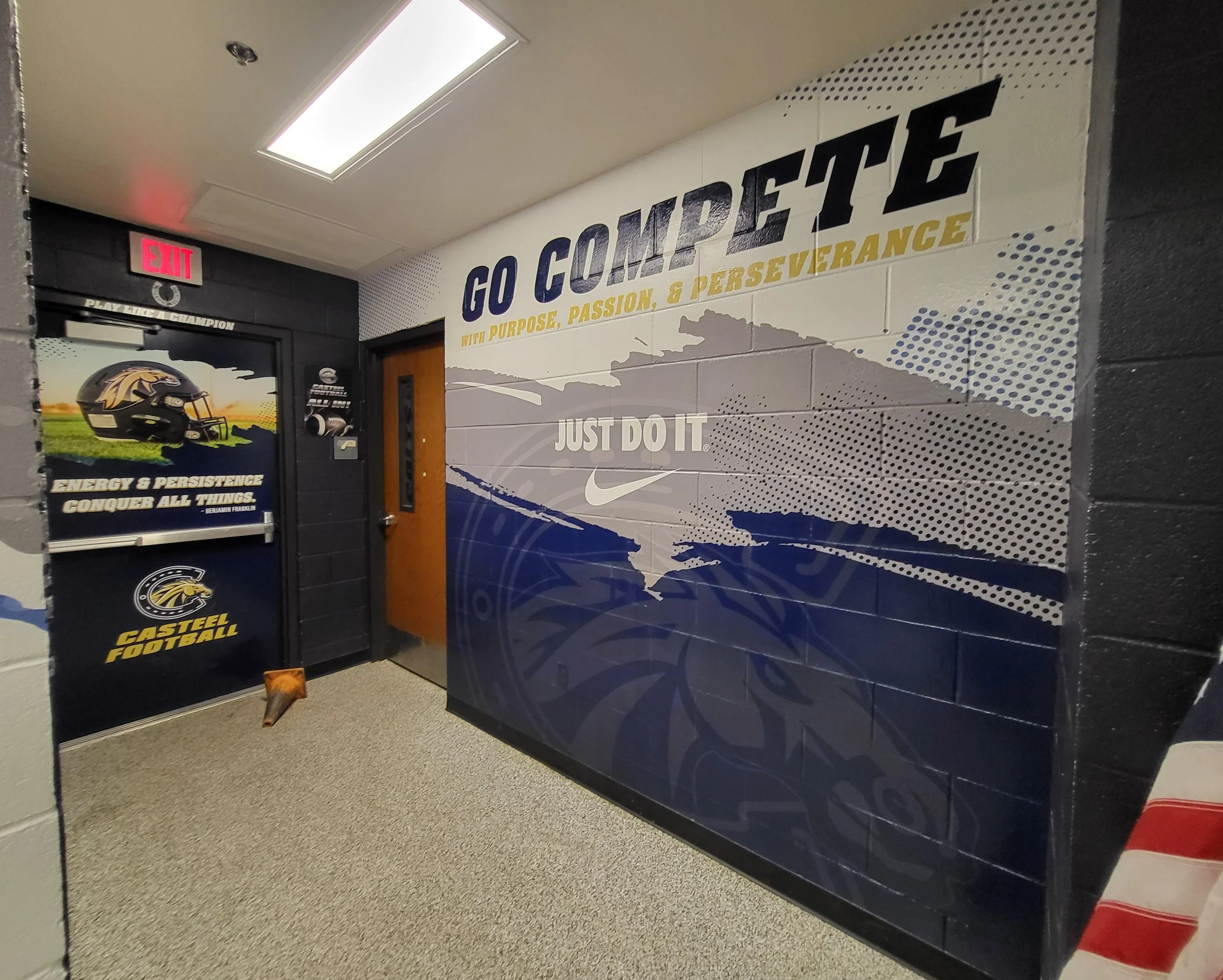 Interior of a school or sports facility hallway with motivational slogans on the wall, a black door with a football helmet graphic, and a red exit sign above the door.