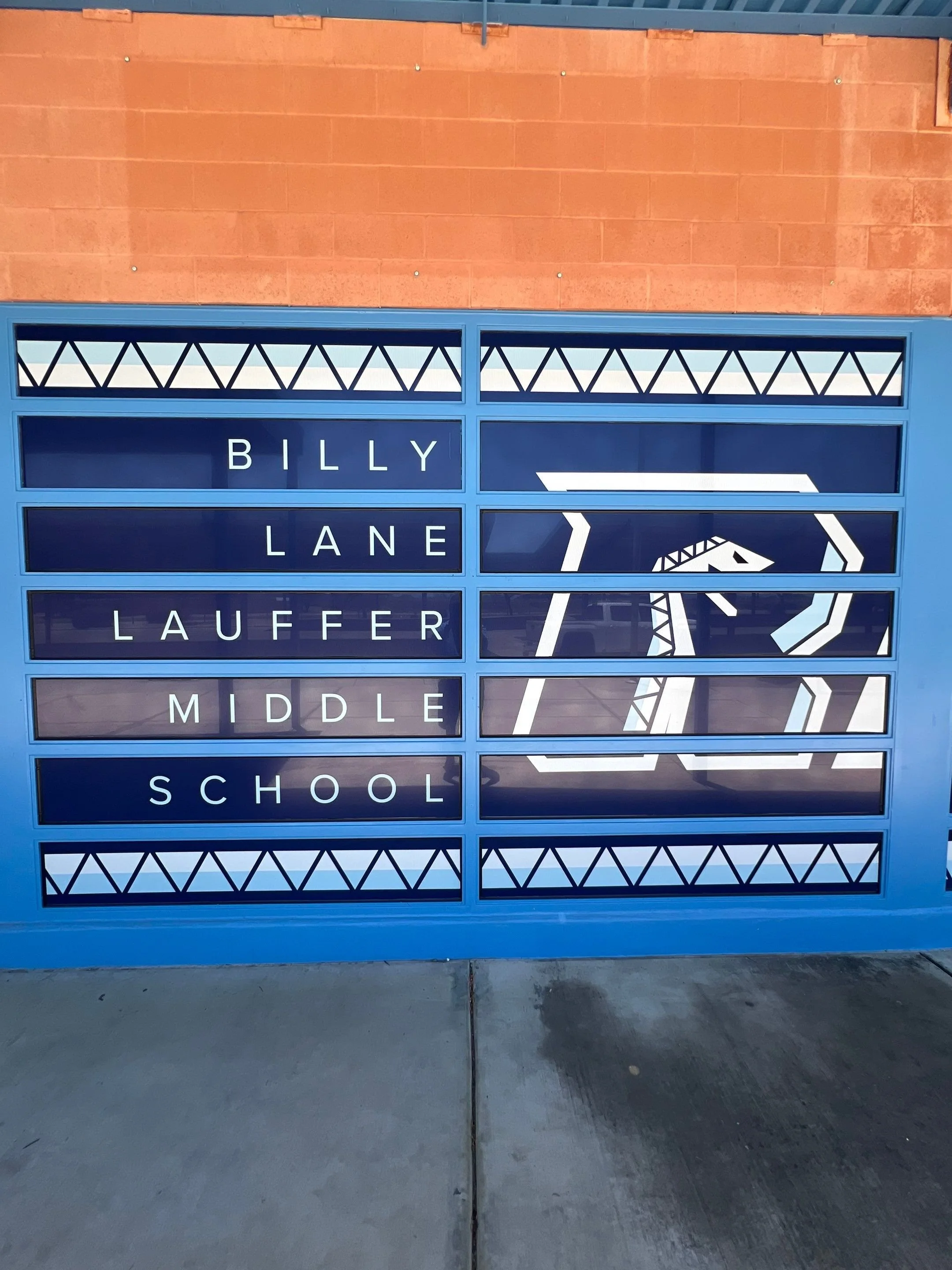 School directional sign with black background and white text listing street names: Billy Lane, Lauffer Middle, and School, with a geometric decorative border and an abstract arrow symbol.