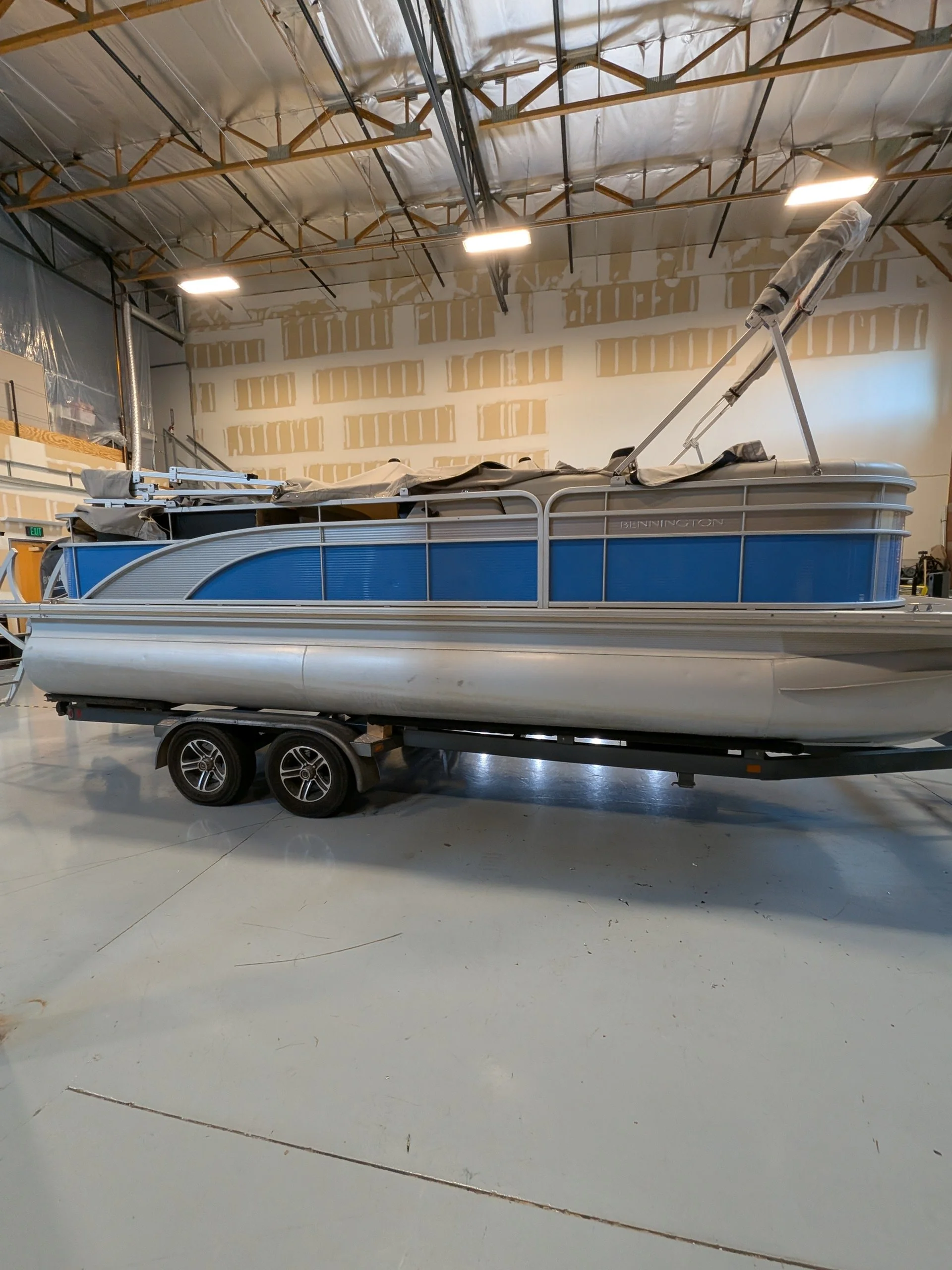 A pontoon boat on a trailer inside a warehouse or workshop. The boat has a blue and gray exterior and a folded canopy or cover on top.