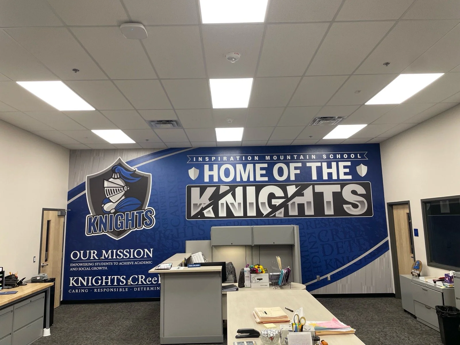 Interior of a school office with a large wall display featuring the Knights mascot, a shield logo, and the text 'Home of the Knights' and 'Our Mission' along with a list of values. Desks, office supplies, and a TV are visible.