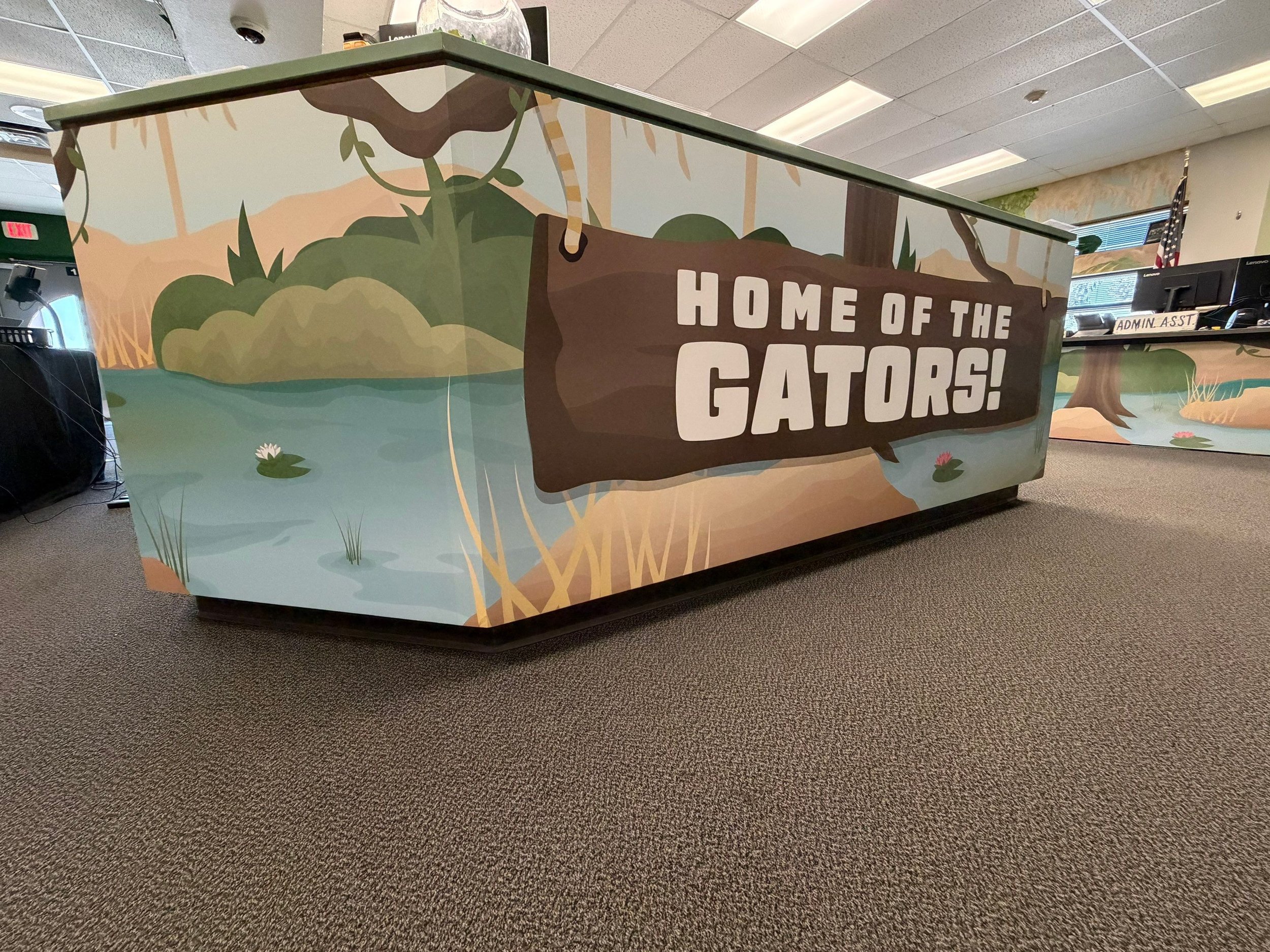 Decorative wooden sign with the text "Home of the Gators!" in large white letters, set against a background featuring a nature scene with trees, water, and hills.