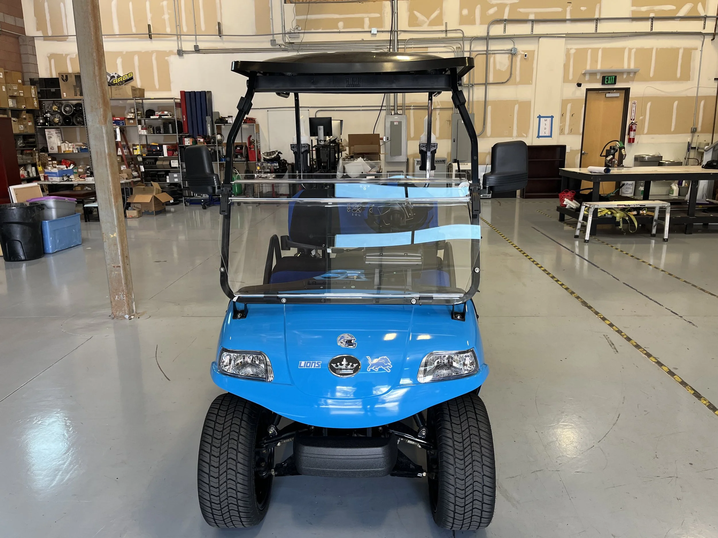 Front view of a blue golf cart with a clear windshield and black roof in a workshop or storage area.