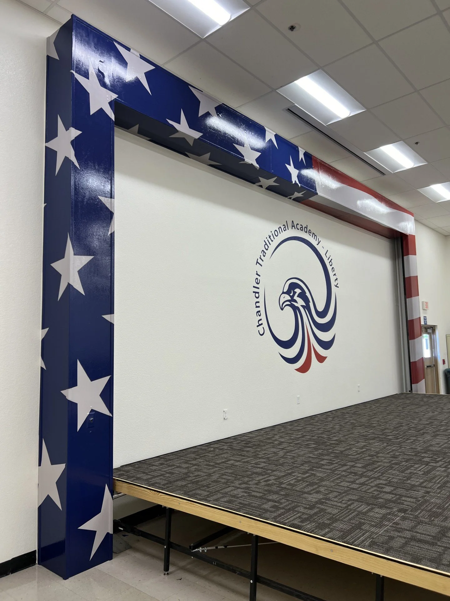Stage with patriotic decorations including a blue pillar with white stars and a wall with a circular logo featuring an eagle and the words "Chandler Traditional Academy - Liberty" and red, white, and blue design elements.