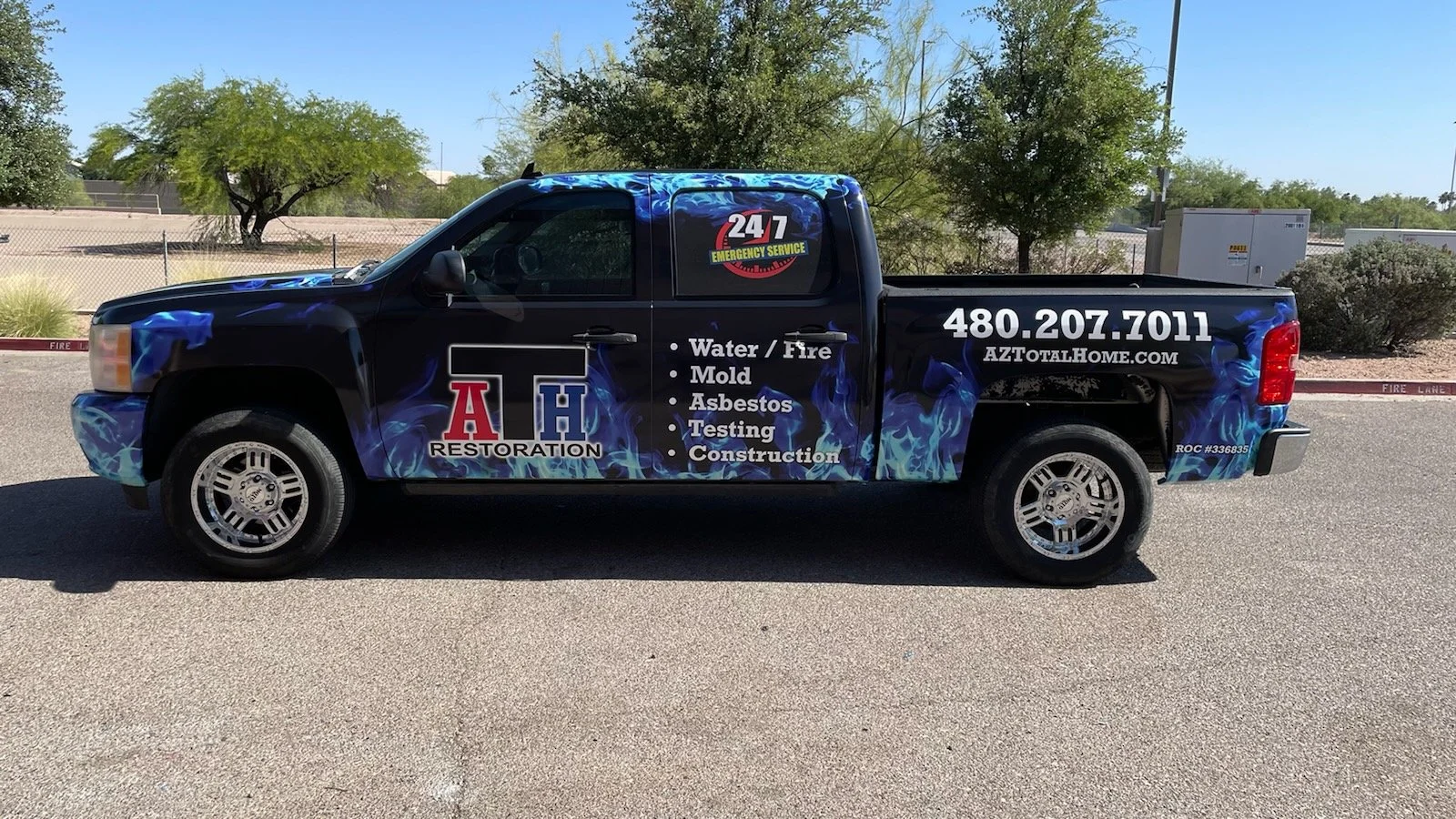 Black pickup truck with advertising for AH Restoration, a water, fire, mold, asbestos, testing, and construction company, parked on a street with trees and a chain-link fence in the background.