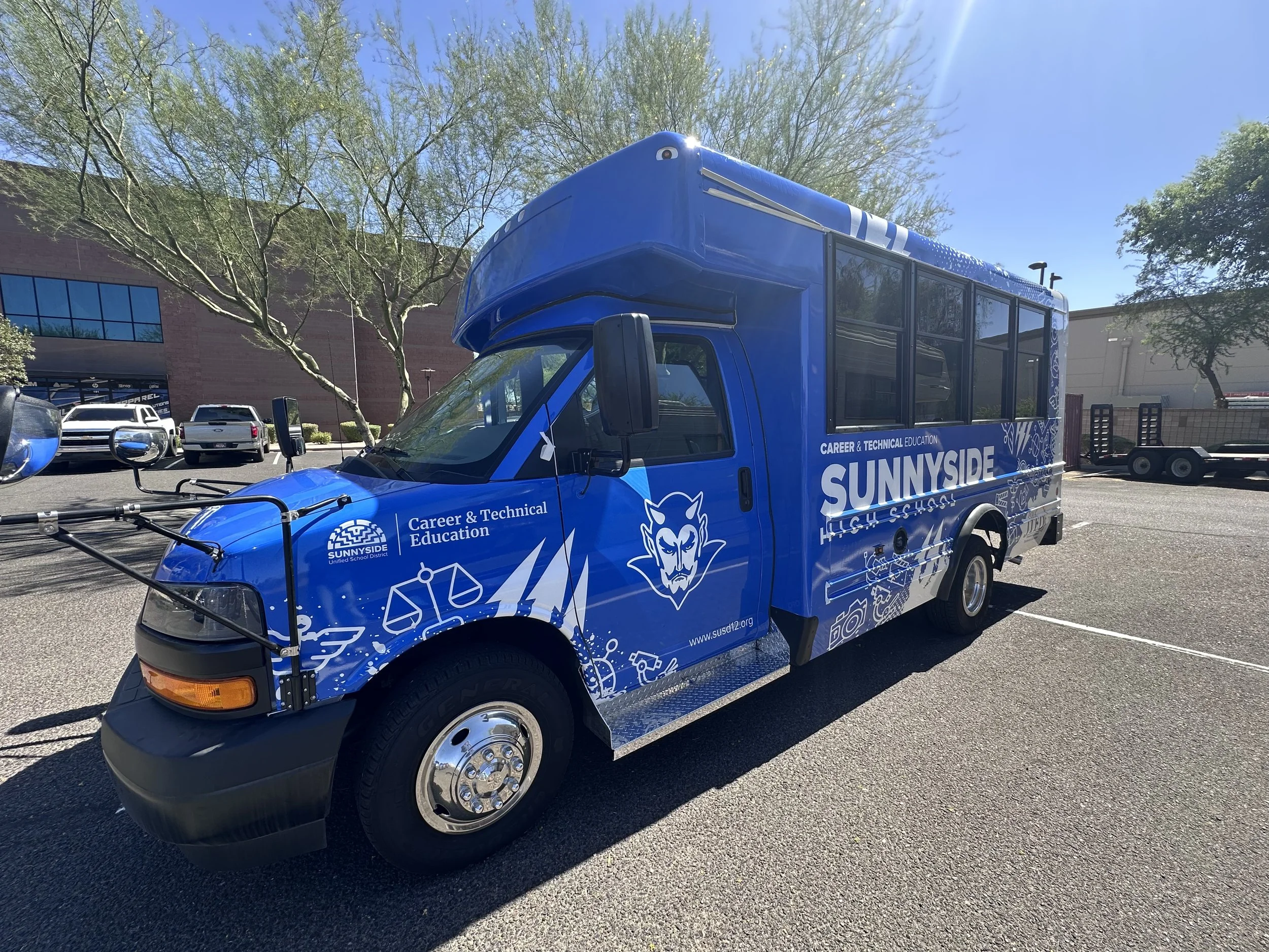 Blue promotional vehicle for Sunnyside, a career and technical education program, parked in a lot with trees and a building in the background.