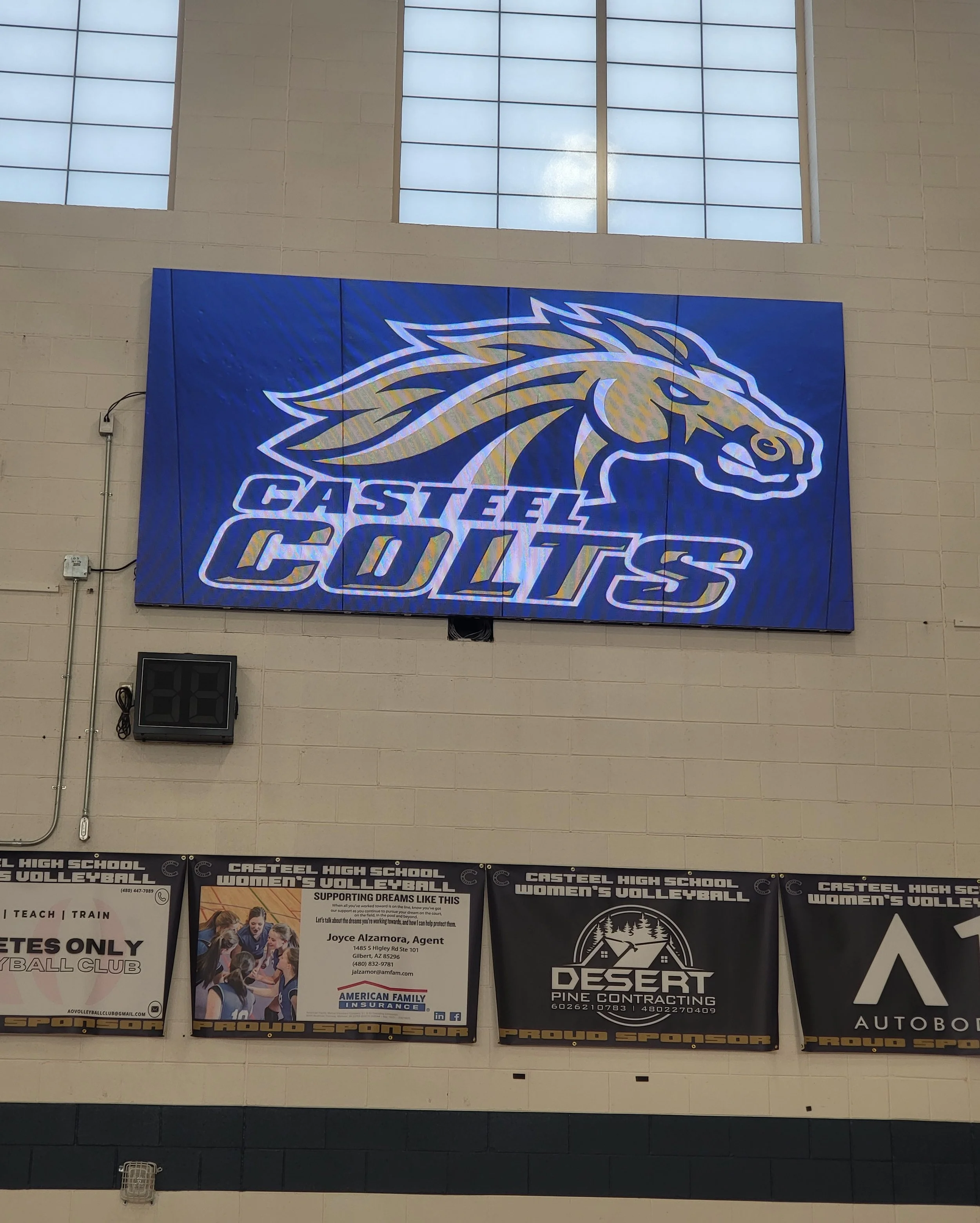 Large indoor digital scoreboard displaying the Castle Colorado logo with a stylized horse head in gold and blue. The background is blue, and the logo includes the text 'CASTLE COLORADO'. The scoreboard is mounted on a beige wall beneath a large window with grid panes.