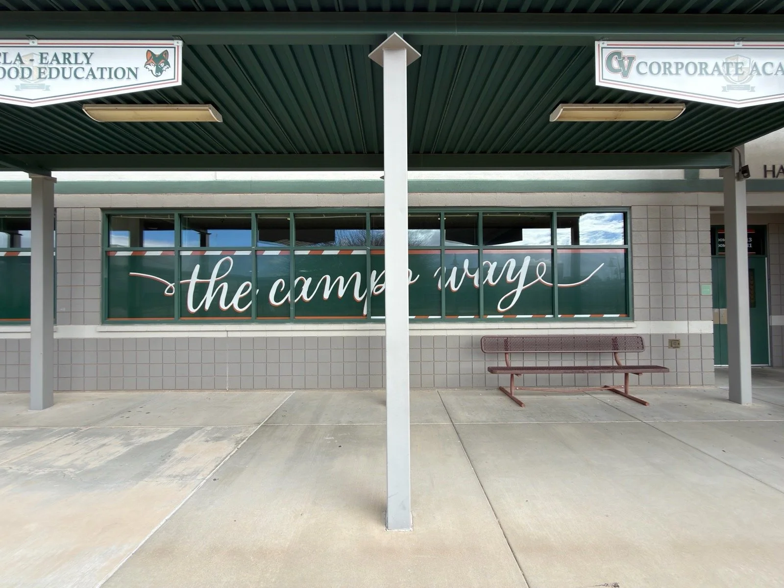 School bus stop with a bench, large windows with the phrase 'the campus way' written on them, and school signs including 'ELA-EARLY FOOD EDUCATION' and 'CV CORPORATE ACADEMY' under a green overhang.