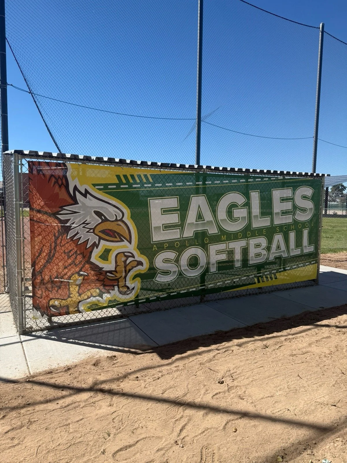 A sign for Apollo Middle School Eagles Softball with a graphic of an eagle, on a fence near a baseball or softball field with blue sky in the background.