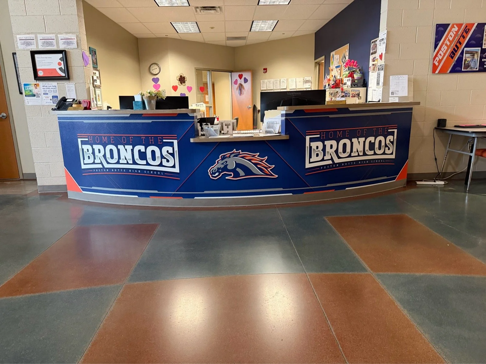 School reception desk with a blue front panel displaying the logo and text 'Home of the Broncos, Poston Butte High School,' decorated with a horse mascot and surrounded by walls with various notices and decorations.