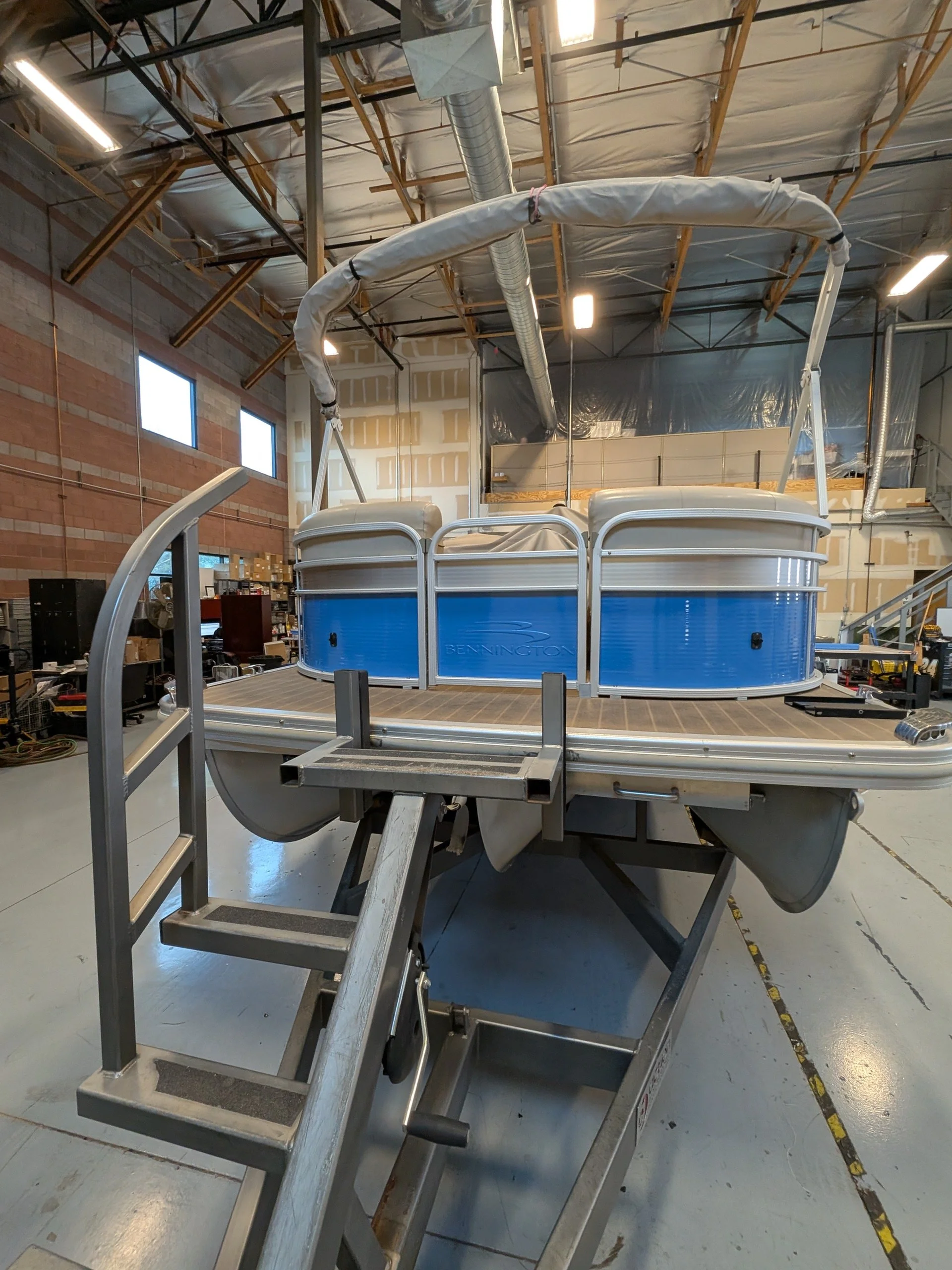 A large, metal pontoon boat with a blue side panel and a walkable deck is inside a warehouse. The boat has a canopy frame and is positioned on the floor, with a mobile ladder in front of it and machinery or supplies around.