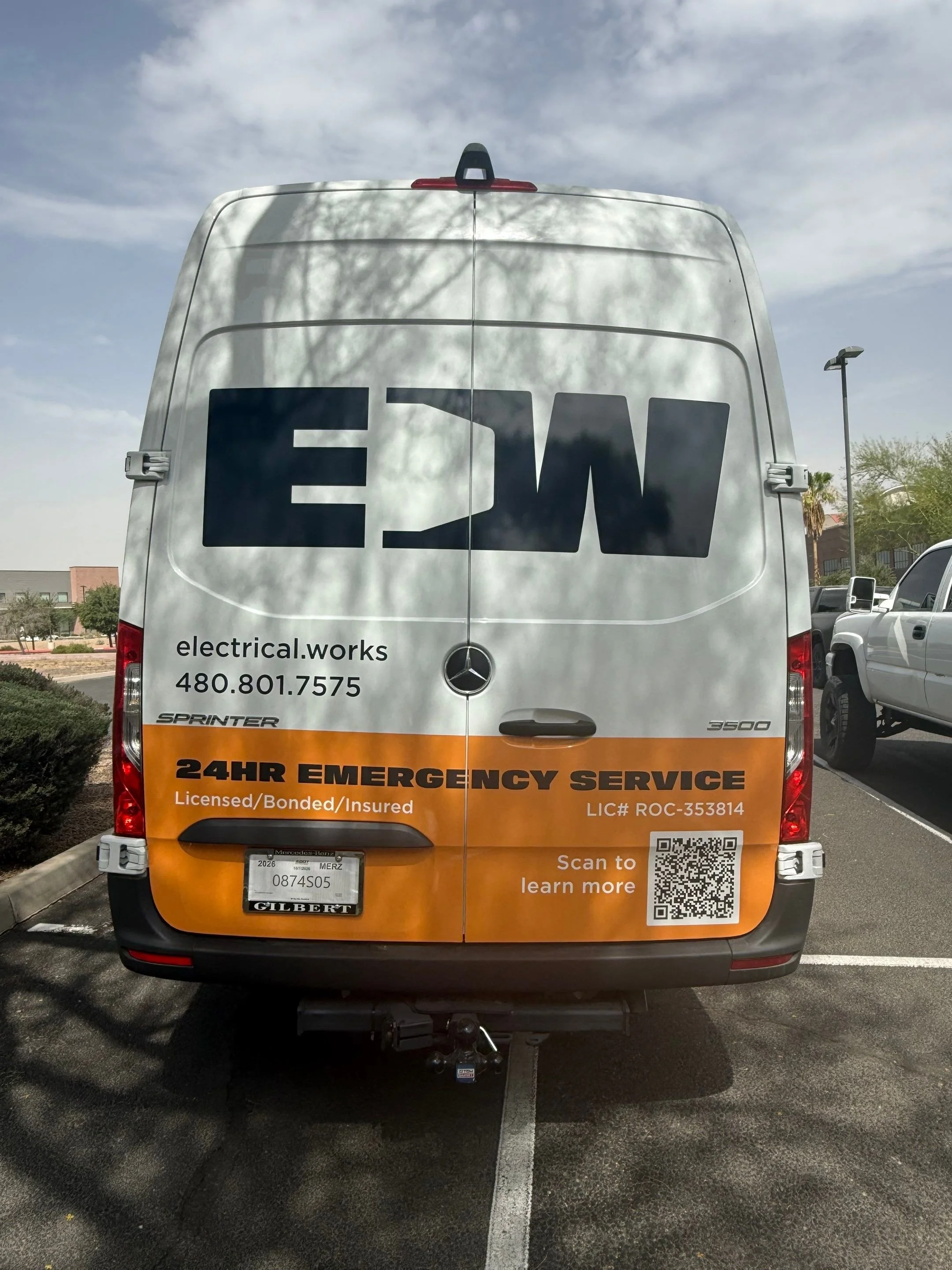 Rear view of a white service van with black and orange graphics, advertising electrical works, emergency service, contact number, and a QR code on the back.