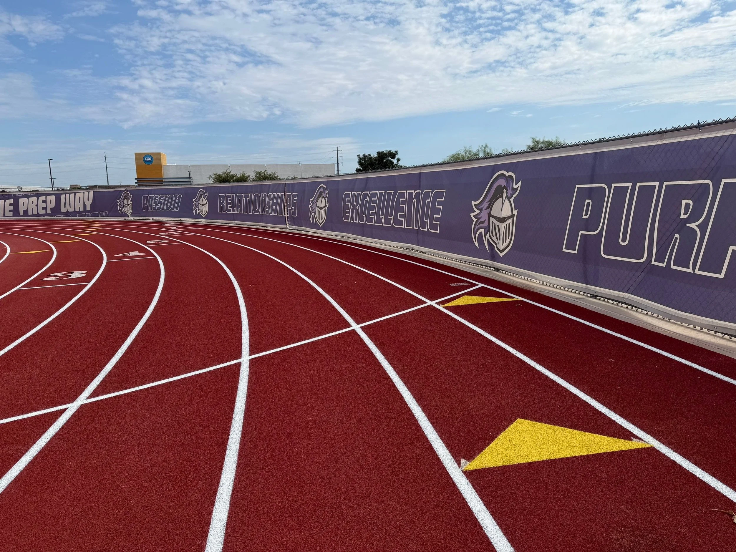 A red running track with white lane lines, surrounded by a purple fence with school-themed graphics and inspiring words like 'PASSION' and 'EXCELLENCE'.