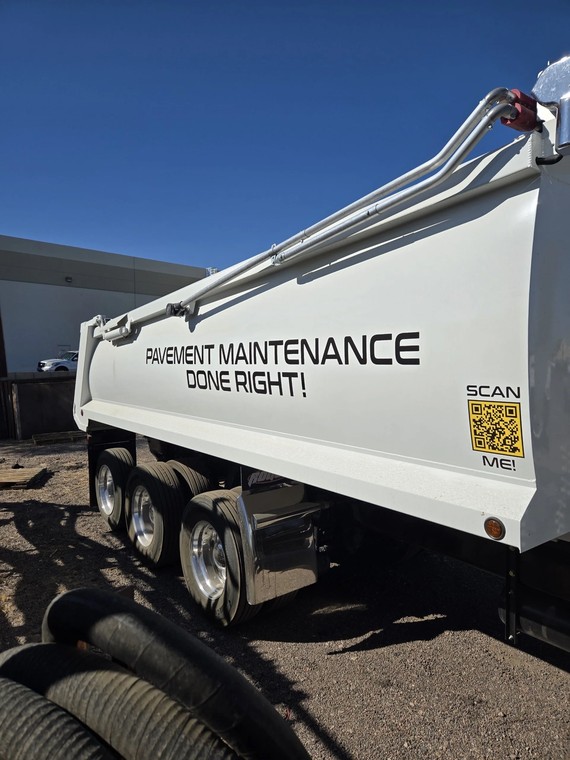 View of a white pavement maintenance truck with black wheels parked outdoors on a sunny day, with a building and a clear blue sky in the background.