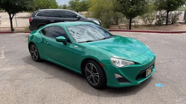 A green two-door sports car parked in a parking lot with a black SUV and trees in the background.