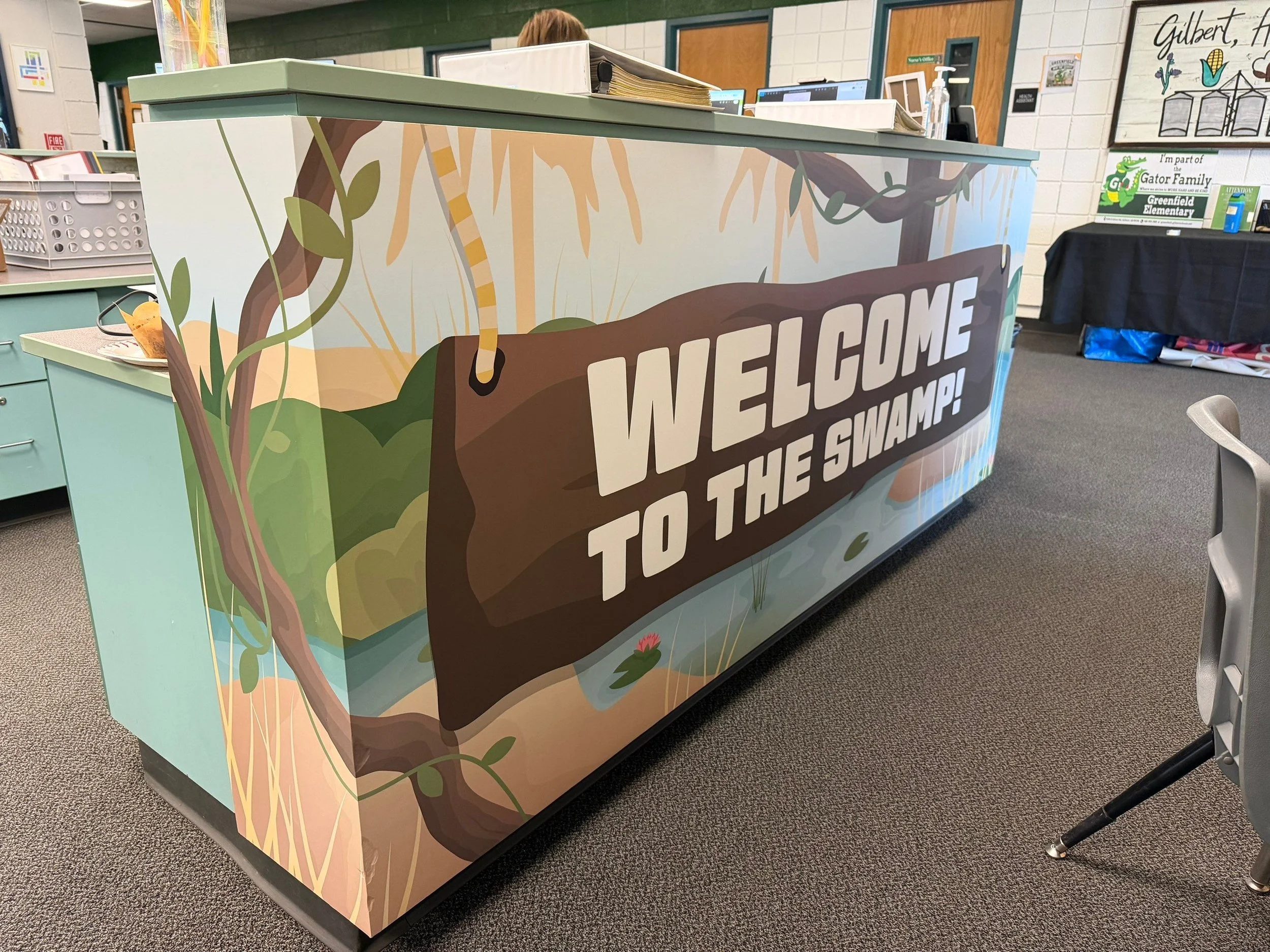 A school reception desk with colorful jungle theme artwork, featuring a large branch and green leaves, with the text "WELCOME TO THE SWAMP!" on the front. In the background, there are desks, computers, and classroom posters in a school setting.