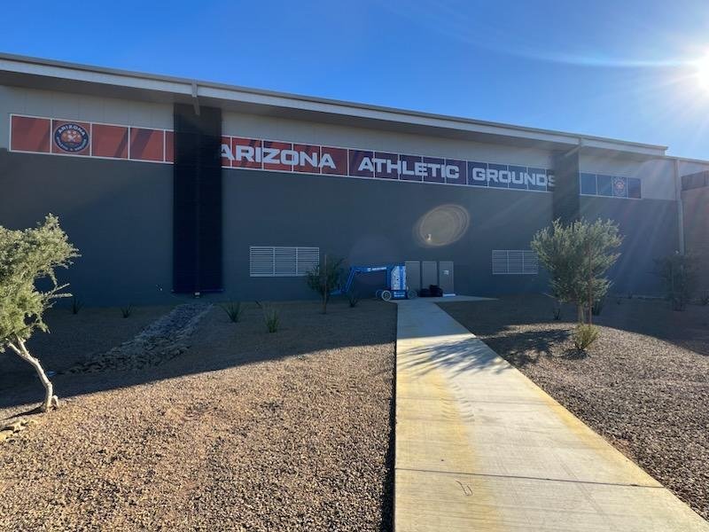 Exterior of Arizona Athletic Grounds with a clear blue sky and the sun shining, a sidewalk leading to the building, desert landscaping, small trees, and some equipment near the wall.