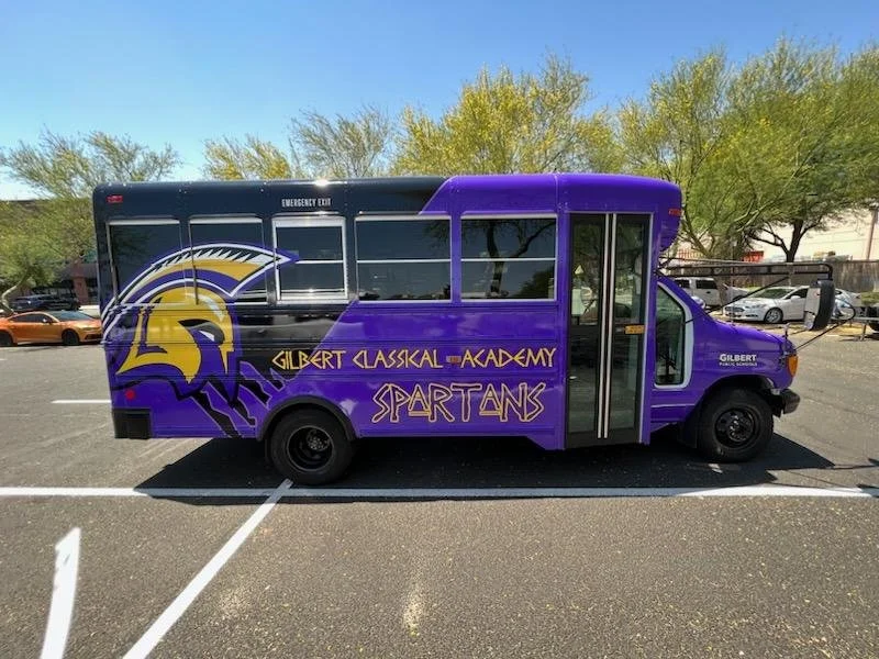 School bus painted in purple with school logo and text for Gilbert Classical Academy Spartans, parked in a parking lot with trees and cars in the background.