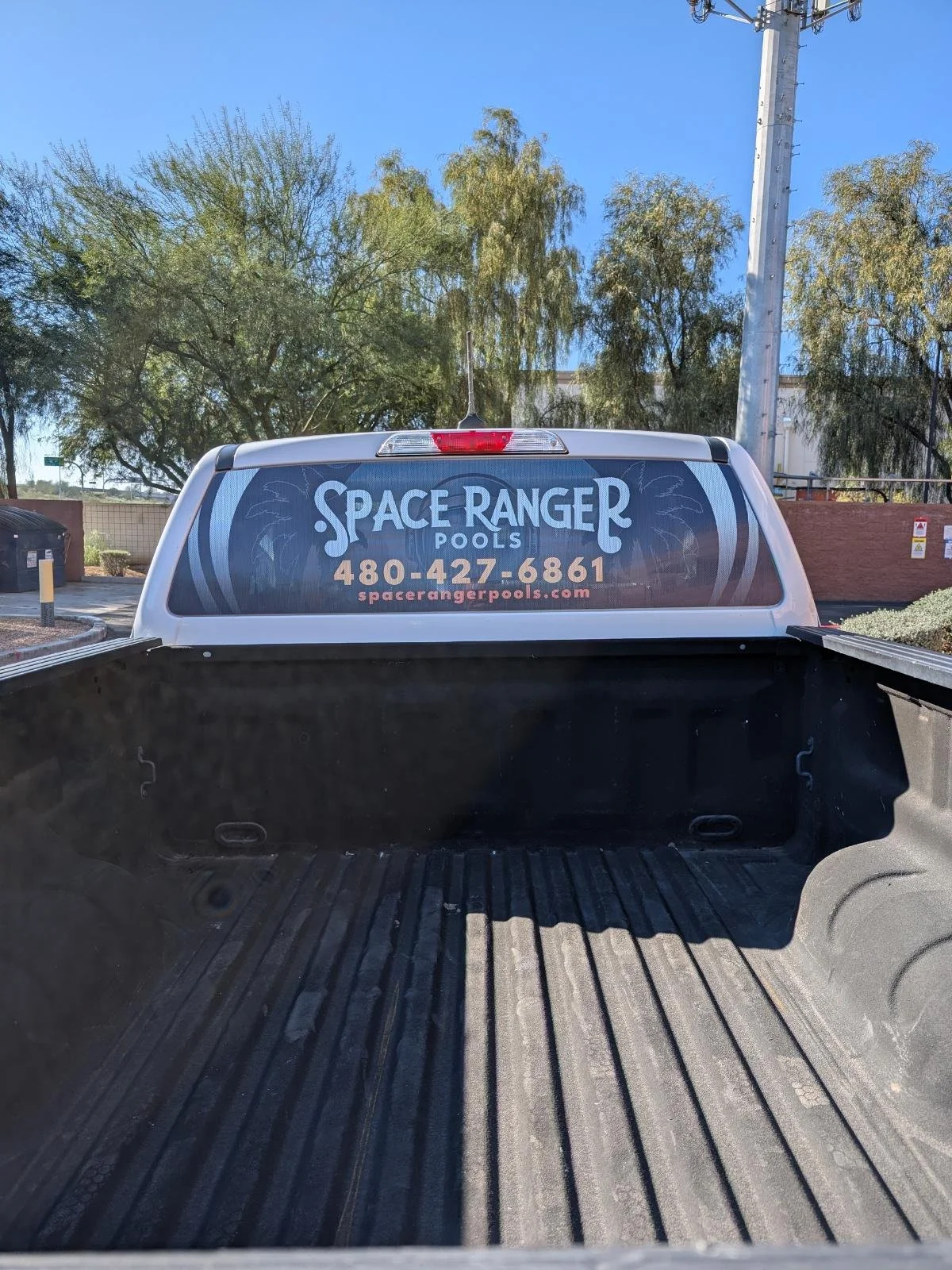 The back of a pickup truck with an advertisement for Space Ranger Pools, including a phone number and website, with trees and a utility pole in the background.