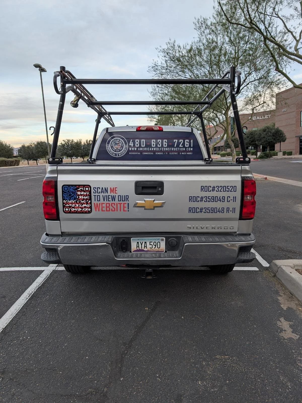 Silver Chevrolet Silverado pickup truck in a parking lot with advertisements and contact information on the tailgate, QR code, and a website URL. The truck has a construction rack on top and Arizona license plate AYA 590.
