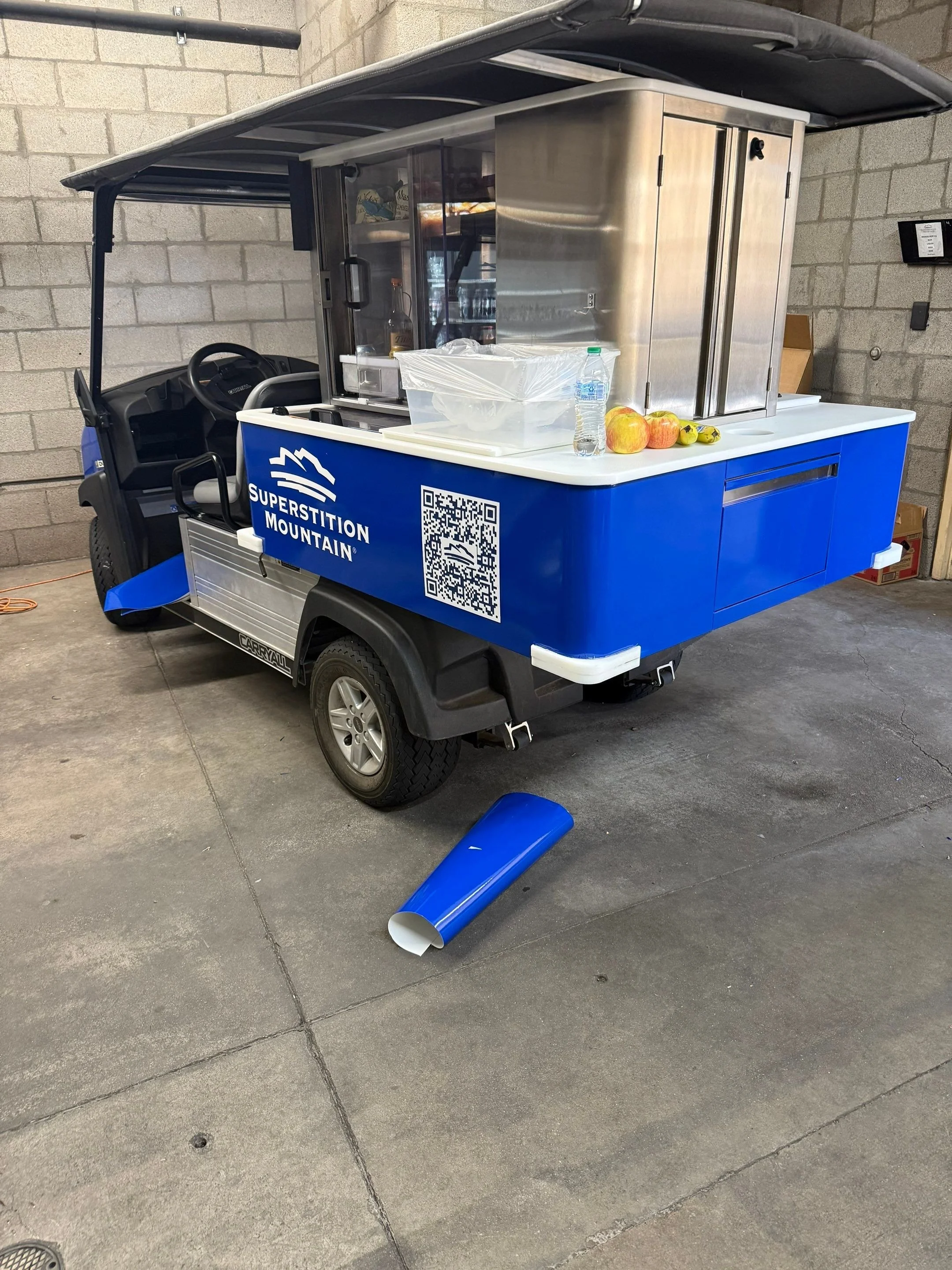 A golf cart with a blue body and a sign that reads 'Superstition Mountain' on the side. The cart is parked on a concrete floor inside a building with a brick wall. The cart has a shaded roof, a steering wheel, and some items on the white counter, inc