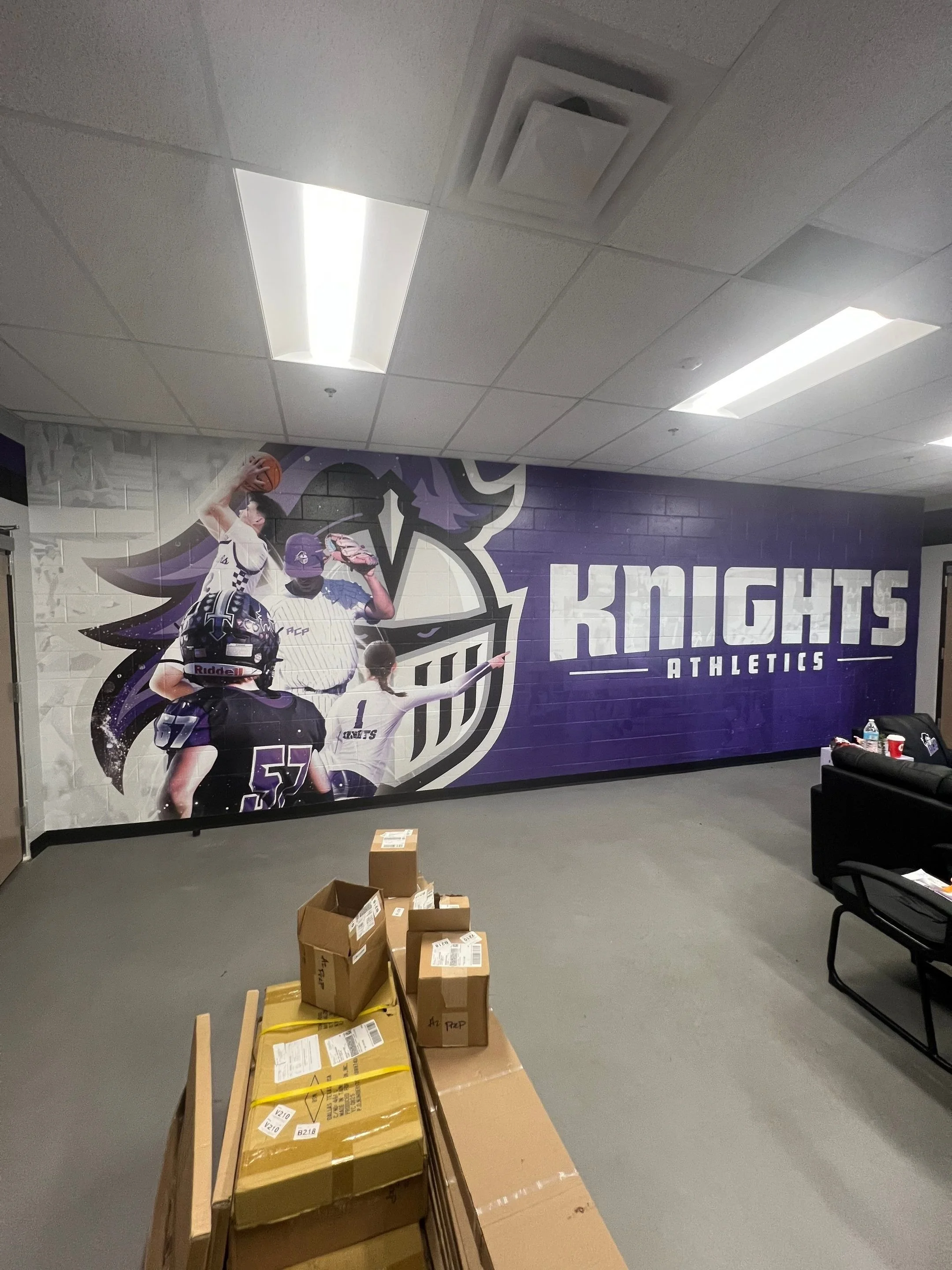 Indoor hallway with a purple and white wall mural of athletes, boxes and furniture in the foreground, ceiling lights, and air vents.