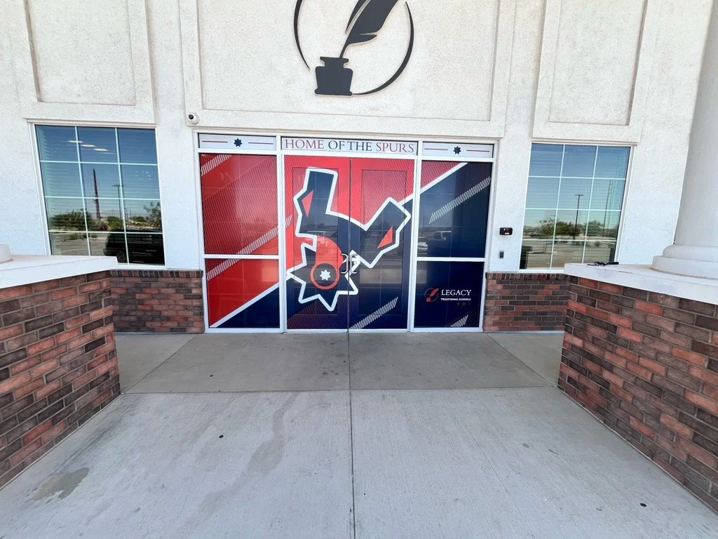 Entrance doors with a decal of a black and white worker or construction silhouette, a red, white, and blue color scheme, and the text "Home of the Spurs" at the top, located outside a building with brick and white facade.