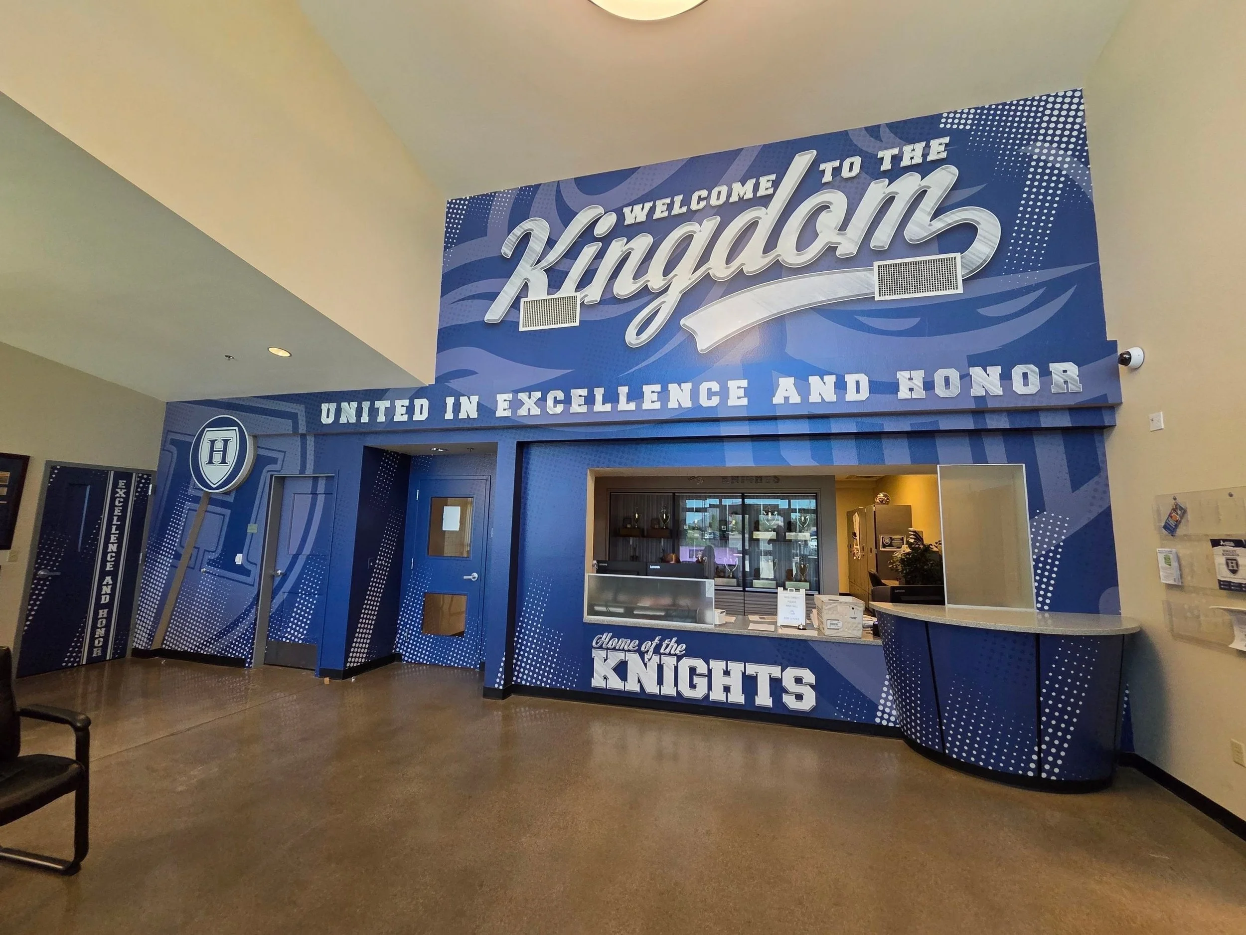 Indoor entrance with a blue wall featuring the large words 'Welcome to the Knights' and the phrase 'United in Excellence and Honor' beneath it, as well as a reception counter and trophies displayed behind glass.