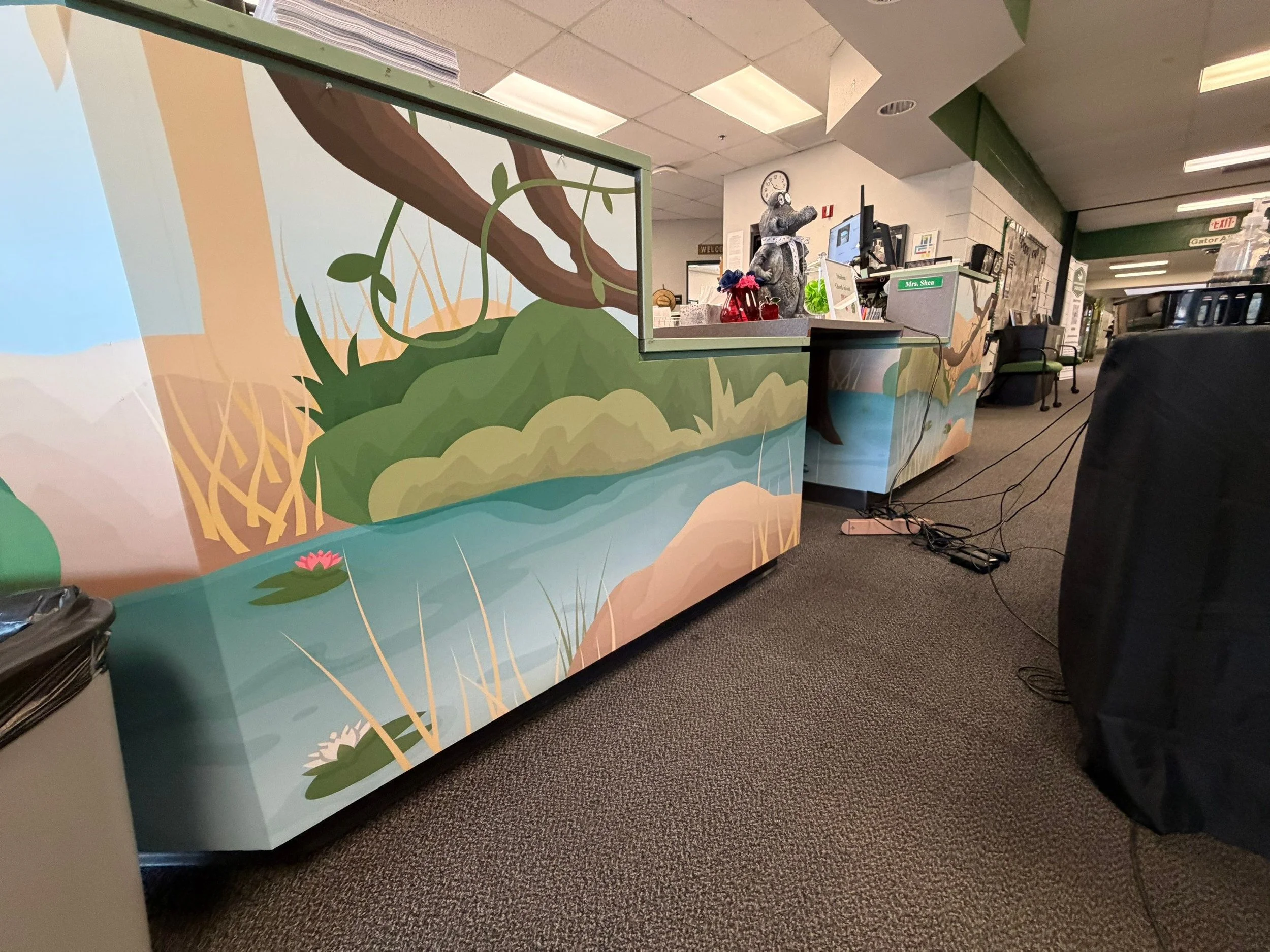 Inside a building at a reception desk with a colorful nature-themed mural of trees and plants on the front, a small statue of a hippopotamus dressed in clothes, and a computer monitor. The room has a drop ceiling with fluorescent lights and a carpete