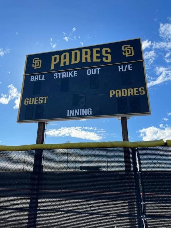 Scoreboard at a baseball field for the San Diego Padres with sections for balls, strikes, outs, hits/errors, guest, Padres, and inning, against a partly cloudy sky background.