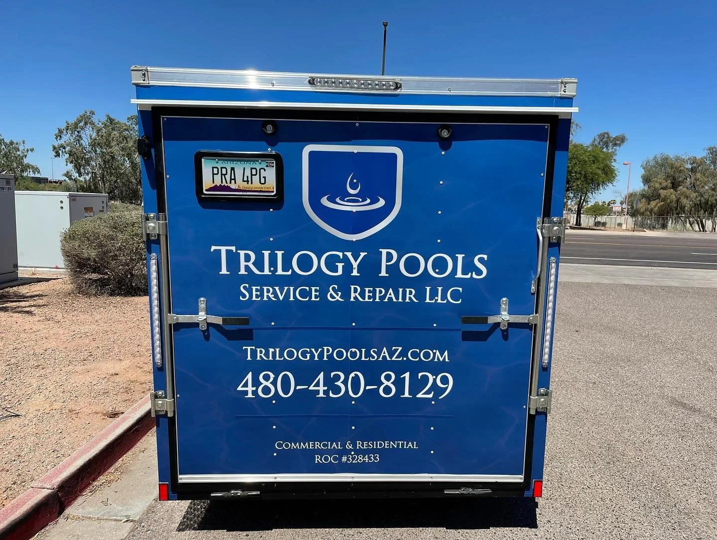 Blue service trailer for Trilogy Pools with company logo, website, phone number, and licensing information parked by the street on a sunny day.