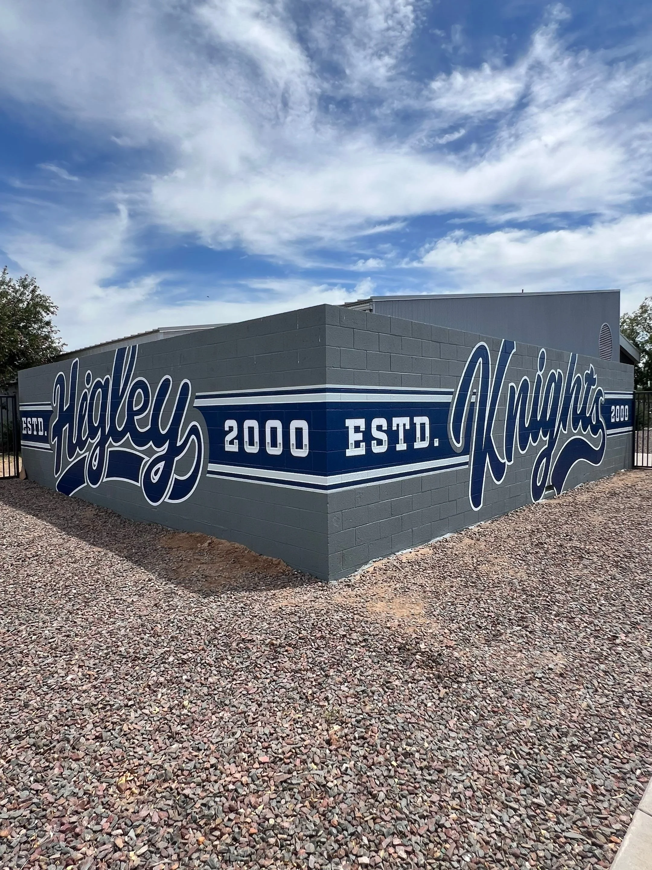 A building with a mural featuring the word 'Highley' in large script, along with 'ESTD. 2000' and 'Knights', painted on a gray brick wall, under a partly cloudy sky.