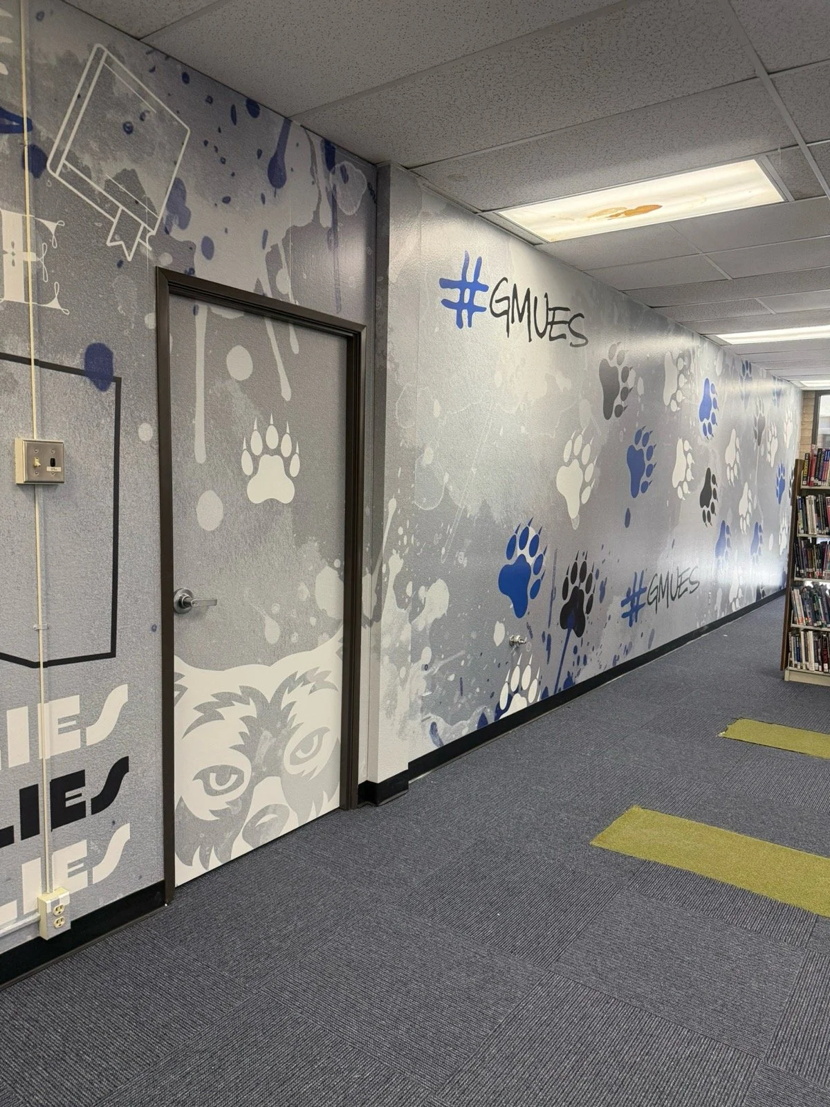 Library hallway wall decorated with paw print patterns and the hashtag #GMUES in black and blue. A closed door with a wolf face illustration at the bottom. Bookshelves on the right side of the image.