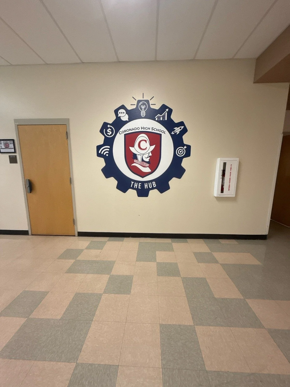 Wall with a large school emblem for Coronado High School, featuring a knight's helmet and gear icons. The emblem reads 'Coronado High School The Hub'. There is a closed door and a white fire alarm on the wall.