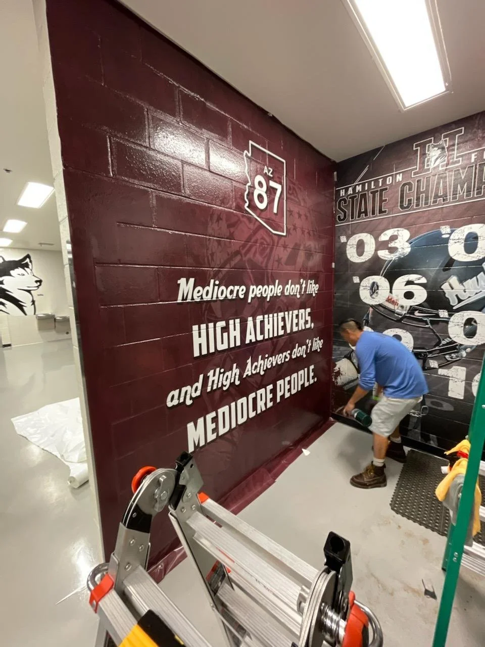 A worker applying a mural to a wall with a quote about mediocrity and achievement, within a room that has a ladder and fluorescent ceiling lights.