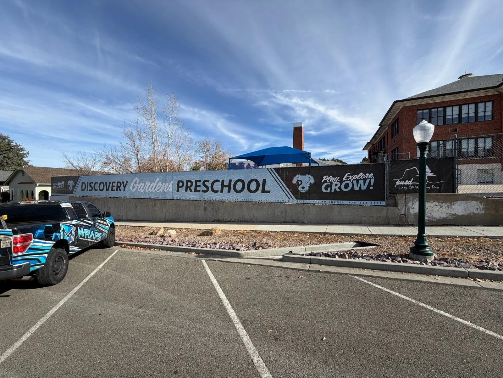 A large banner on a wall promoting Discovery Gardens Preschool, with the slogan 'Play. Explore. Grow!' and contact information, in front of a parking lot with a black vehicle and a street lamp.