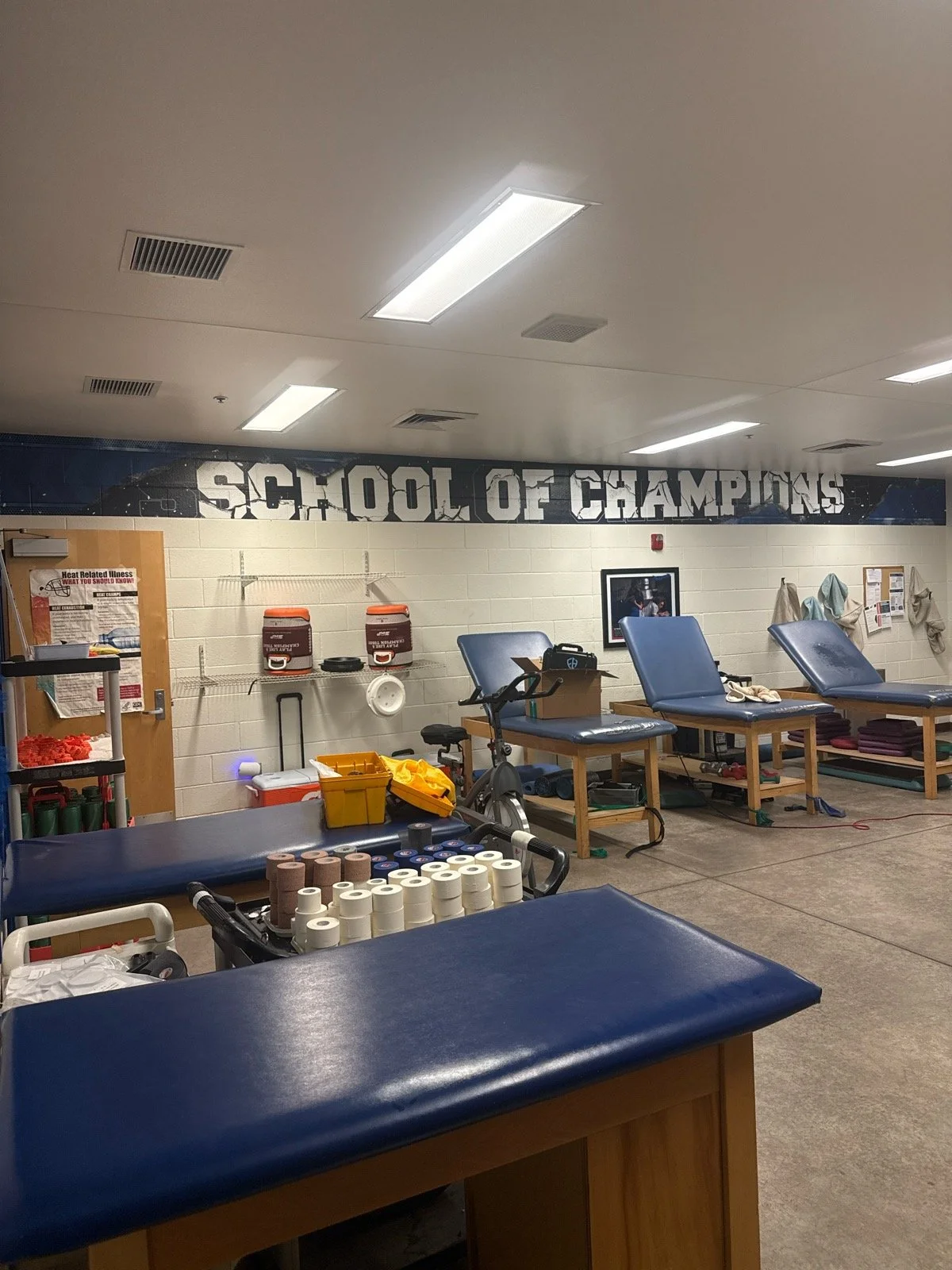 A room in a school gym or sports facility with medical and athletic equipment, including treatment tables, a stationary bike, and supplies, under a large sign reading 'School of Champions.'