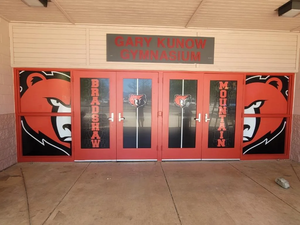 Entrance doors of Gary Kunow Gymnasium with school mascot, a fierce bear, and words 'Bradoshaw' and 'Mountain' printed on the glass.