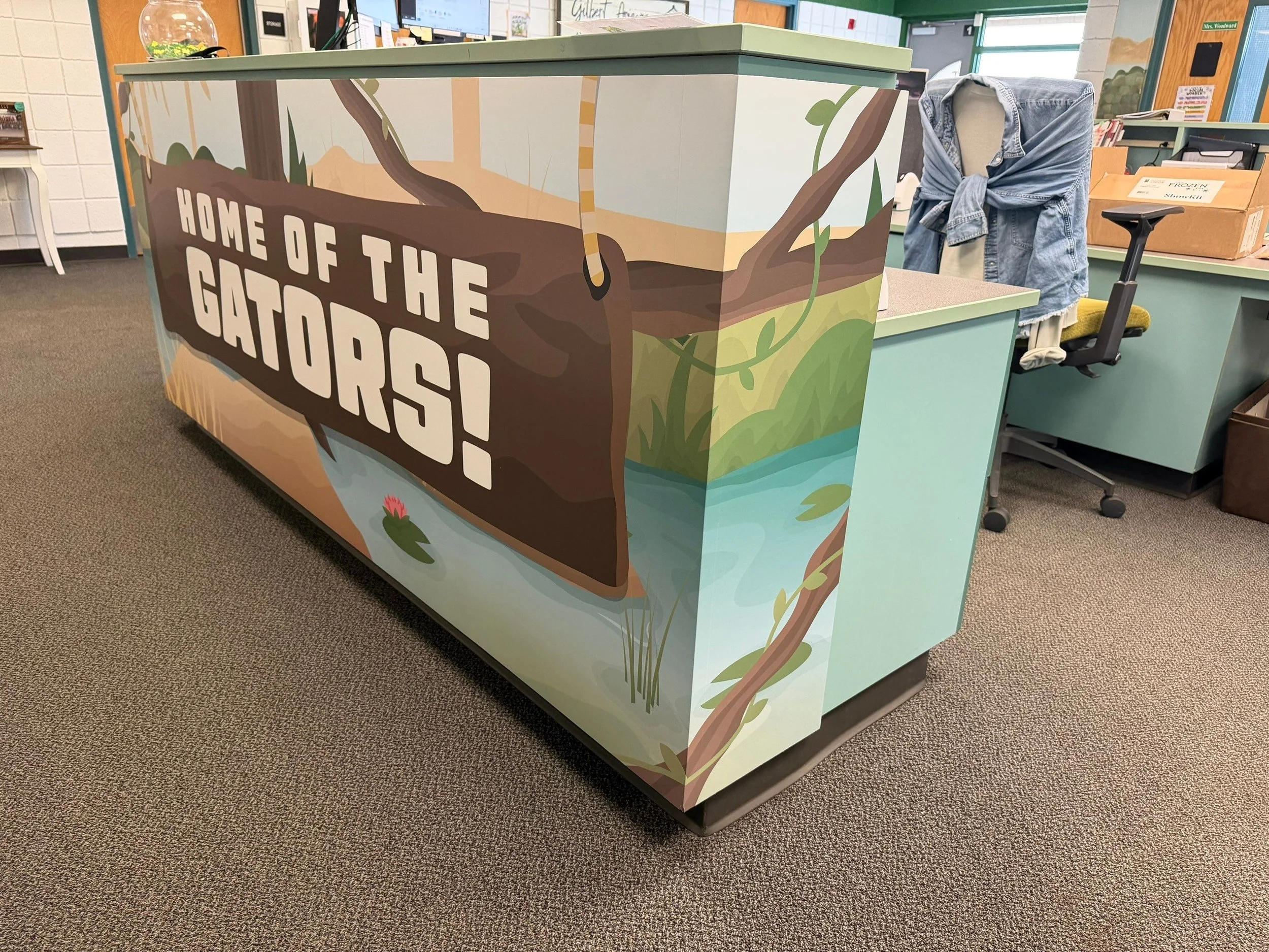 A colorful desk or counter with a nature-themed mural featuring trees, vines, water, and a pink flower, with the words "Home of the Gators" written on it.