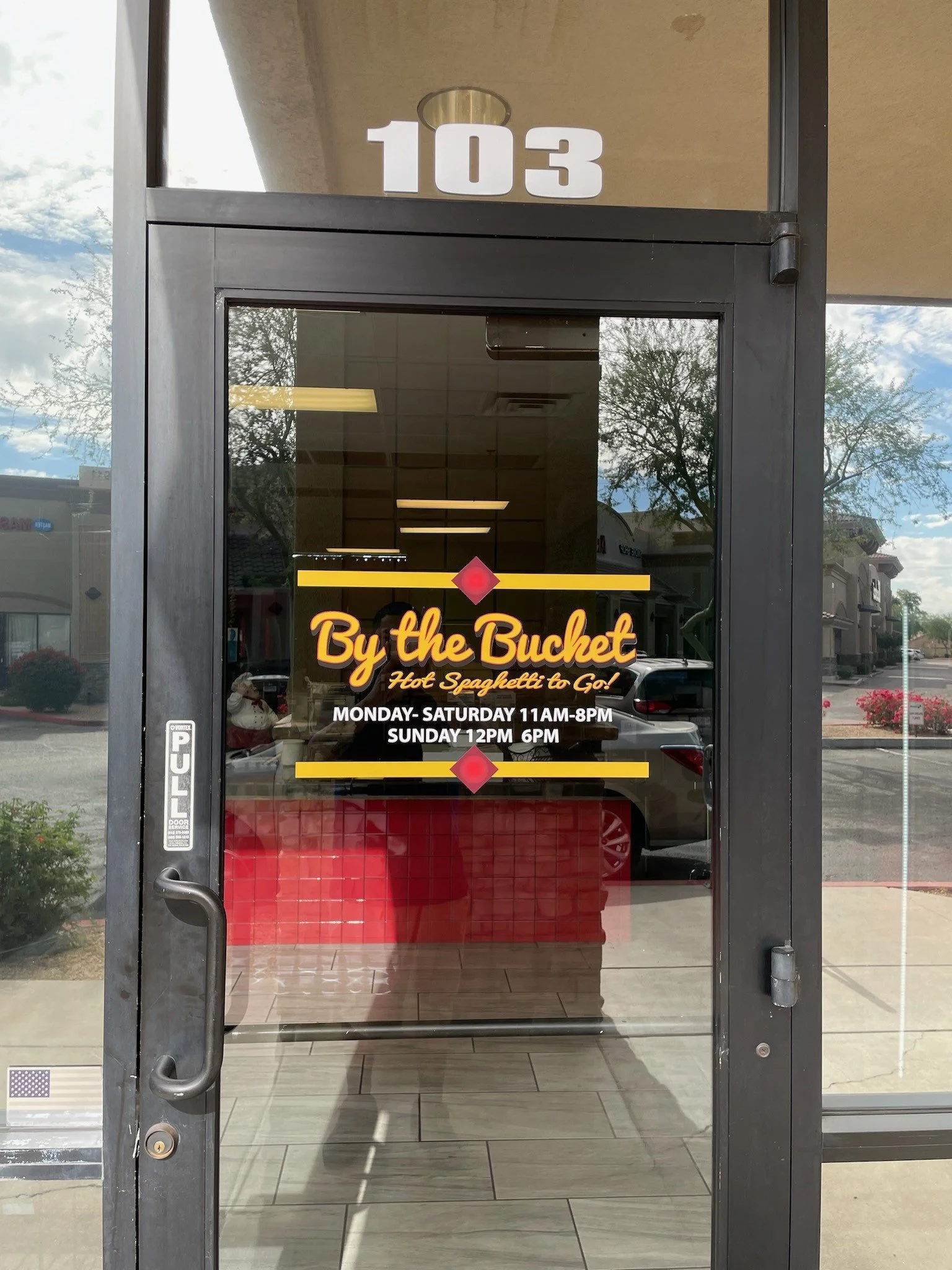 The glass door of a restaurant named 'By the Bucket' with the address number '103' above it. The door has signage including the restaurant's name, slogan 'Hot Spaghetti to Go!', operating hours, and a 'Pull' handle on the left side.