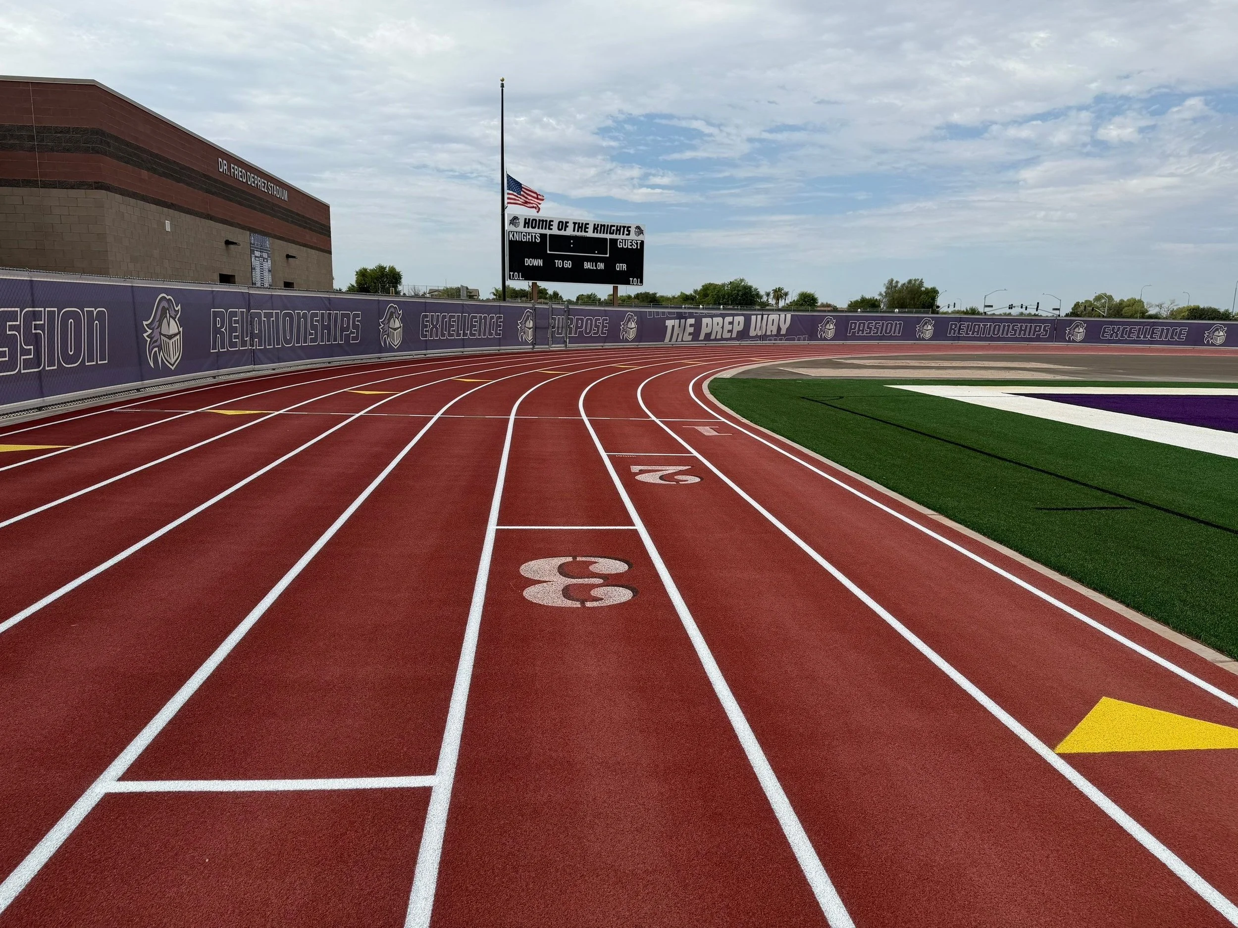 An outdoor running track at Dr. Fred Deprez Stadium with red lanes, a green infield, and a purple fence displaying the words 'Relationships,' 'Excellence,' 'Purpose,' and 'Passion' along with a logo. A scoreboard and American flag are visible in the 