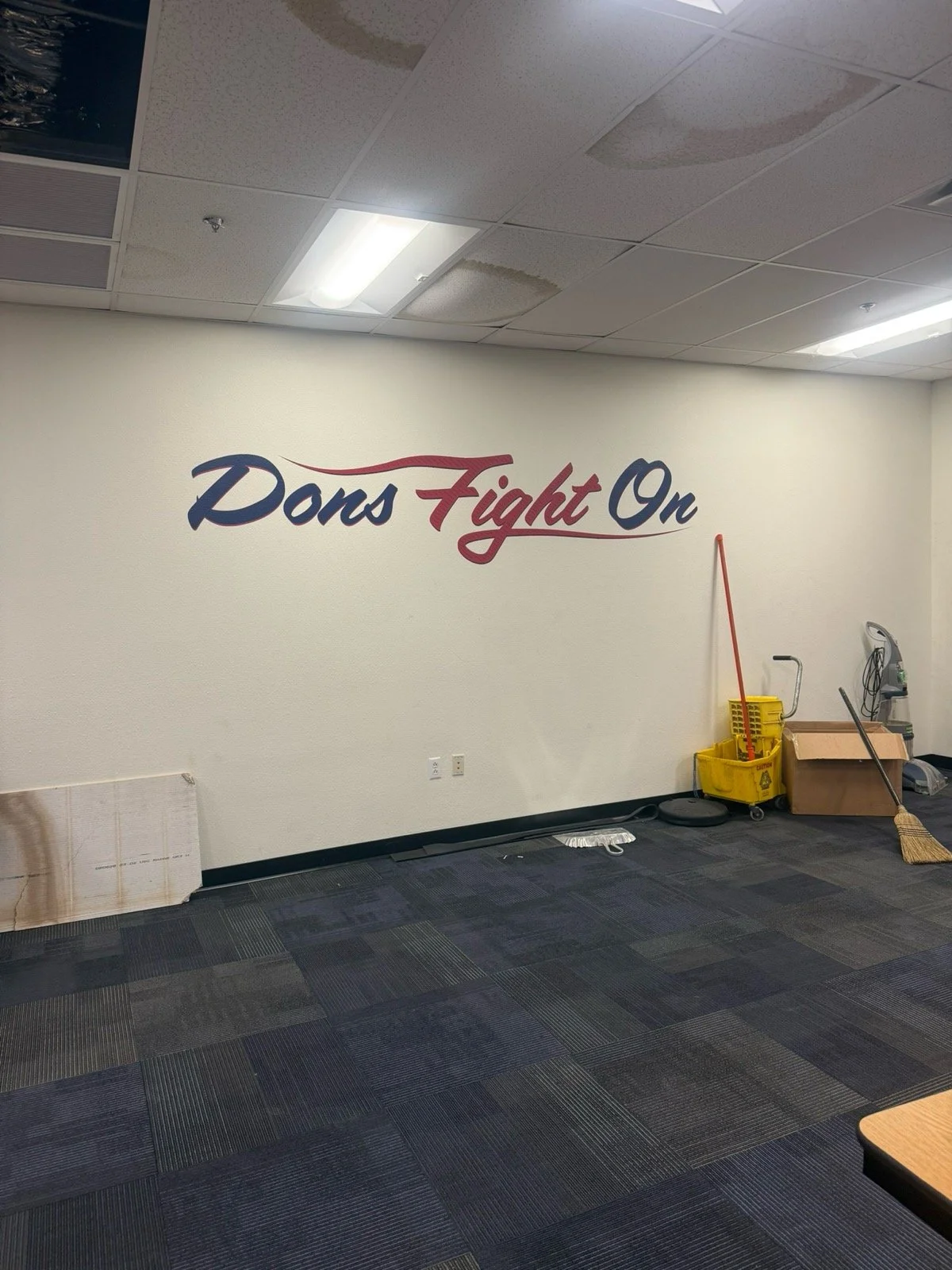 A wall with the phrase 'Dons Fight On' painted in blue and red cursive letters, part of a room with cleaning supplies and equipment on the floor.