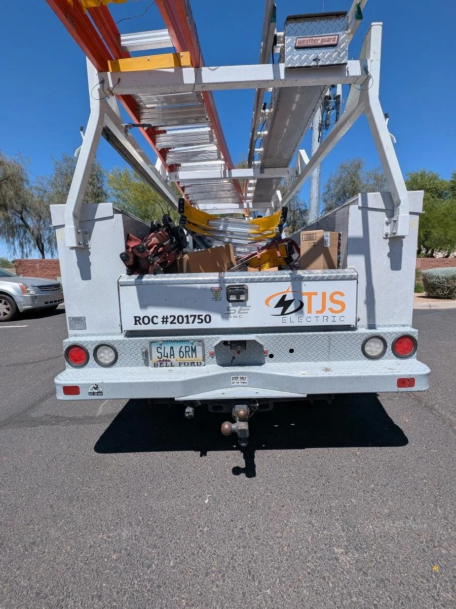 Rear view of a white TJS Electric utility truck with a ladder extended upwards, carrying tools and boxes, in a parking lot on a sunny day.