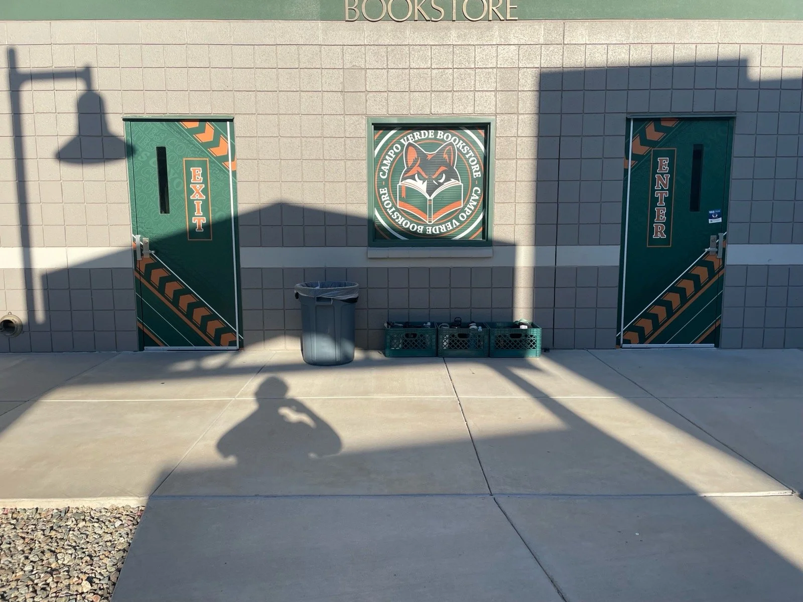 Exterior of a bookstore with two green doors labeled Exit and Enter. There is a mural of a fox reading a book in the center, surrounded by a circular logo that reads 'Camp Verde Bookstore.' A trash can and crates with bottles are in front of the buil